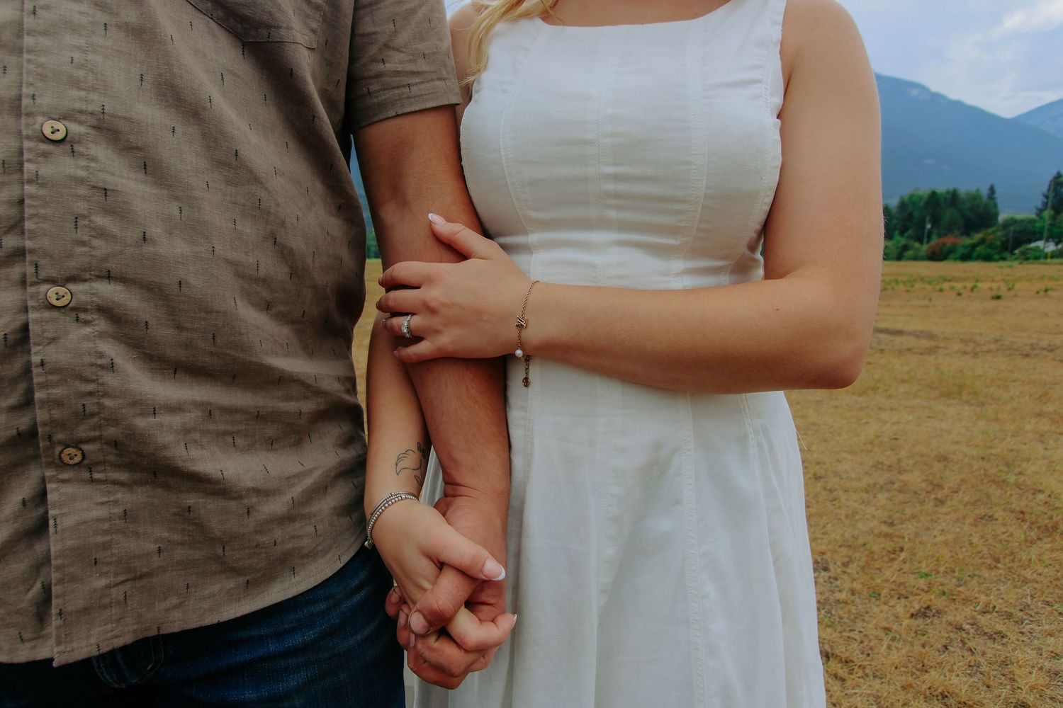 Couple holding hands in a field, woman in white dress with mountains in background.