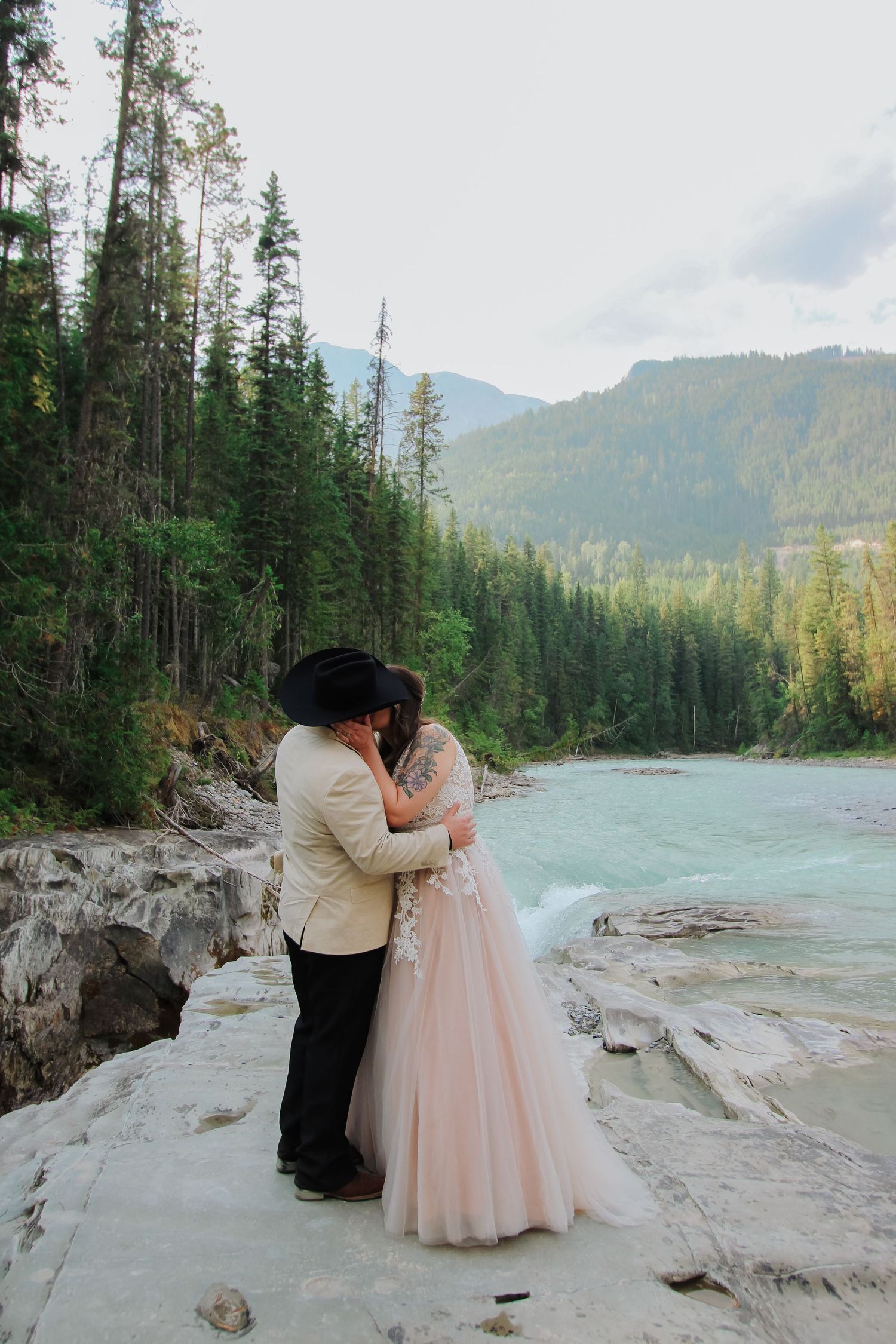 Couple sharing a kiss in wedding attire by a turquoise river with mountain forest backdrop.