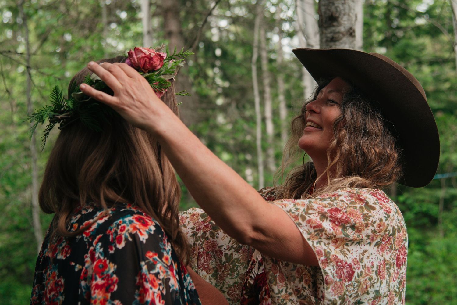 Woman in cowboy hat places floral crown on another woman outdoors in a lush green forest setting.