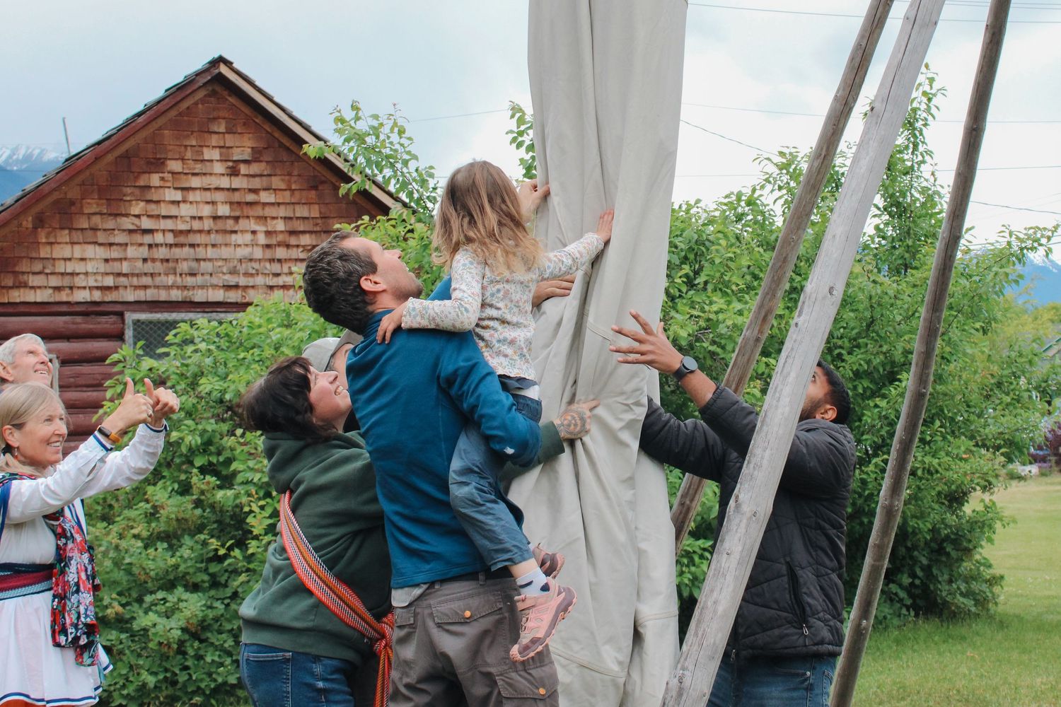Group of people raising a large white canvas tent outdoors near a wooden cabin surrounded by green trees.