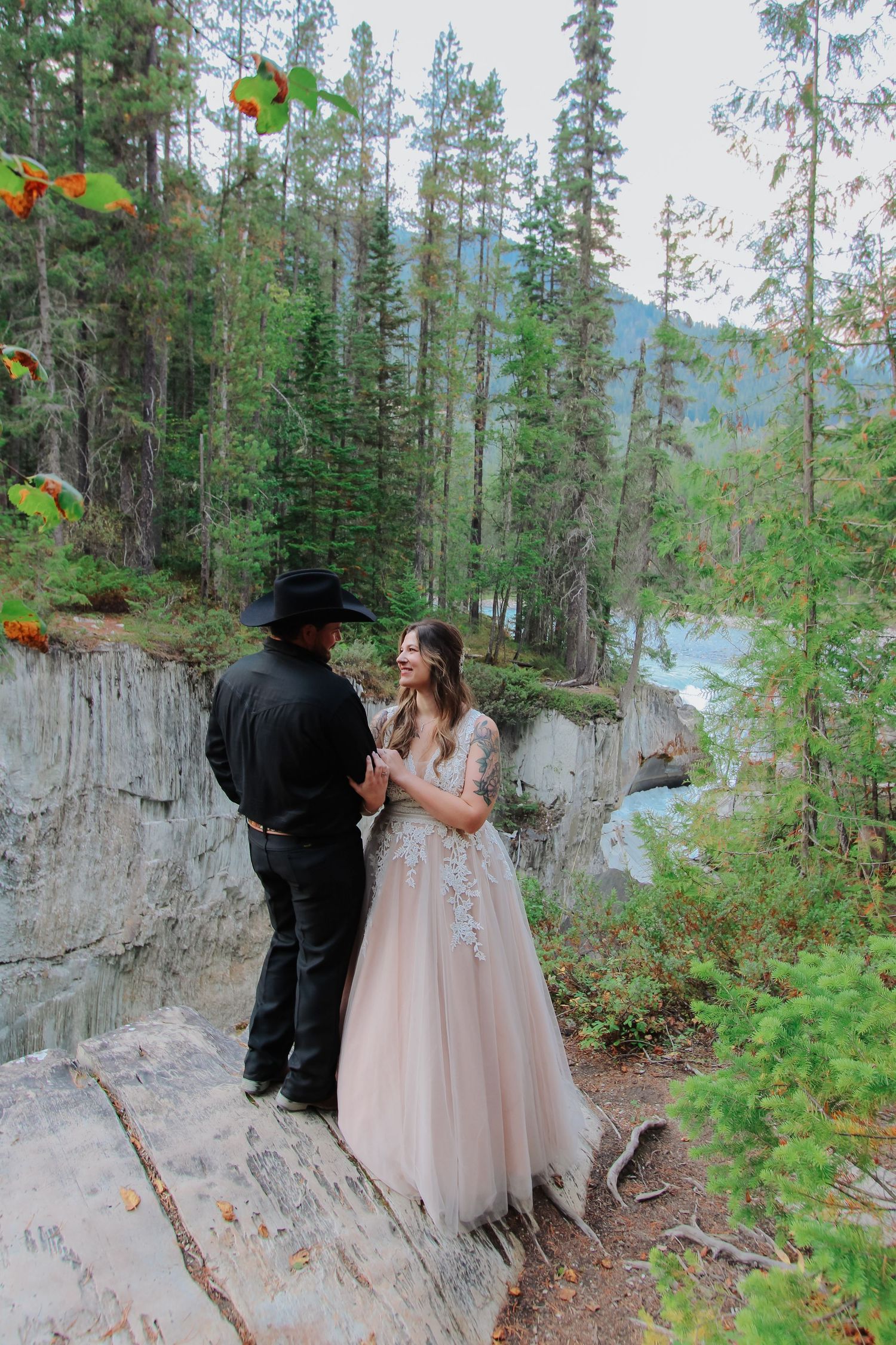 Couple in wedding attire sharing vows on rocky cliff edge surrounded by pine forest and river in Canadian Rockies.