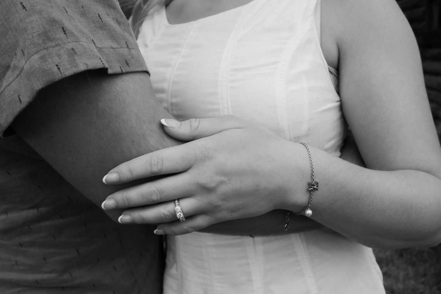 Black and white photo of a couple embracing, woman wearing an engagement ring and bracelet in a white dress.