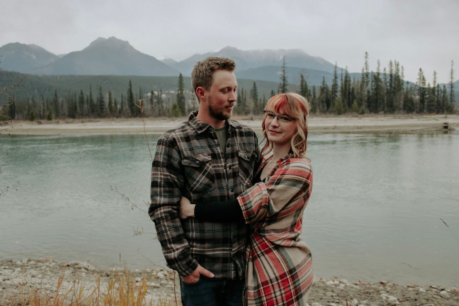 A couple embraces by a scenic mountain lake on an overcast day.