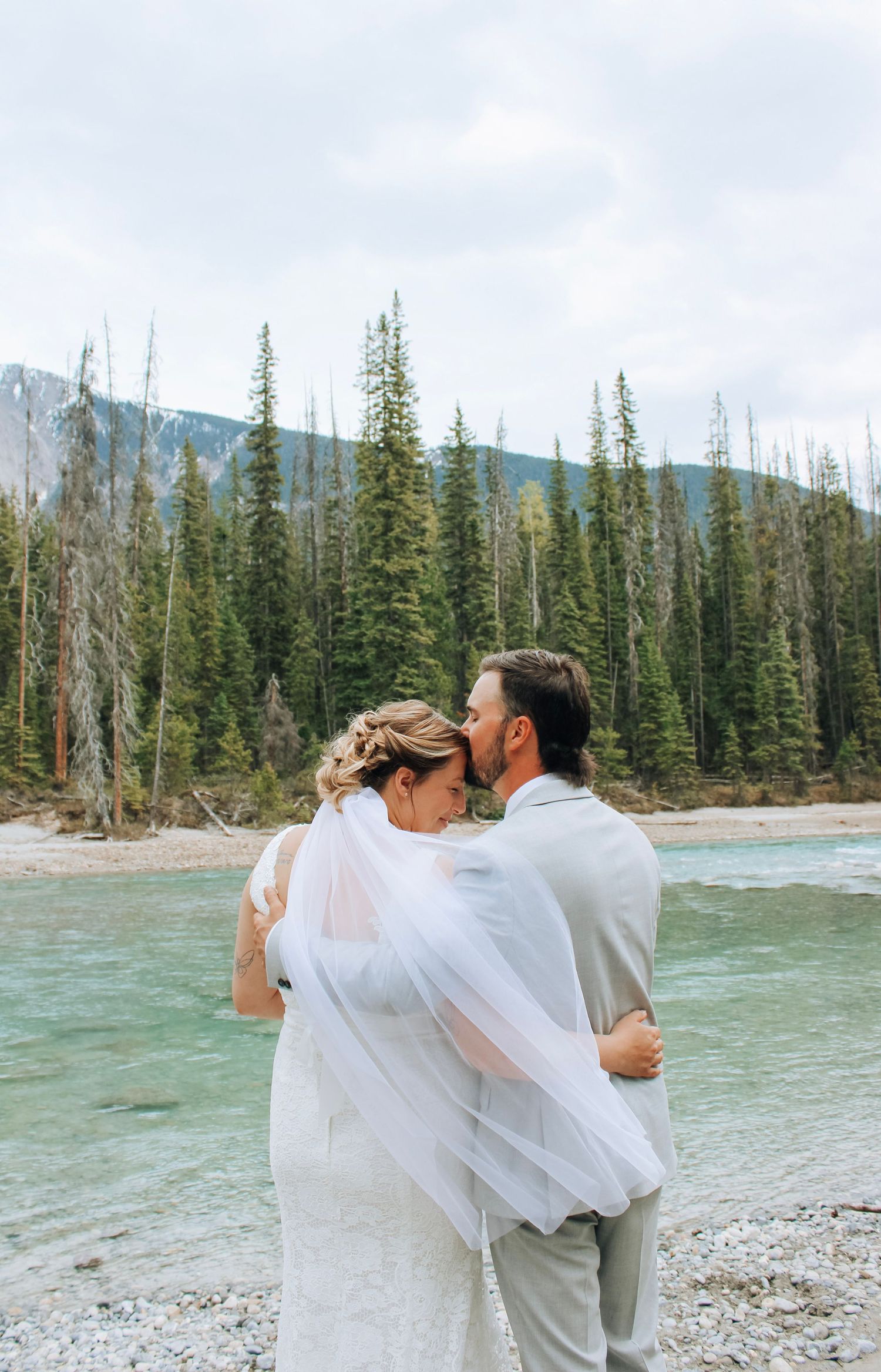 Romantic wedding portraits by a turquoise mountain lake with evergreen forest backdrop.