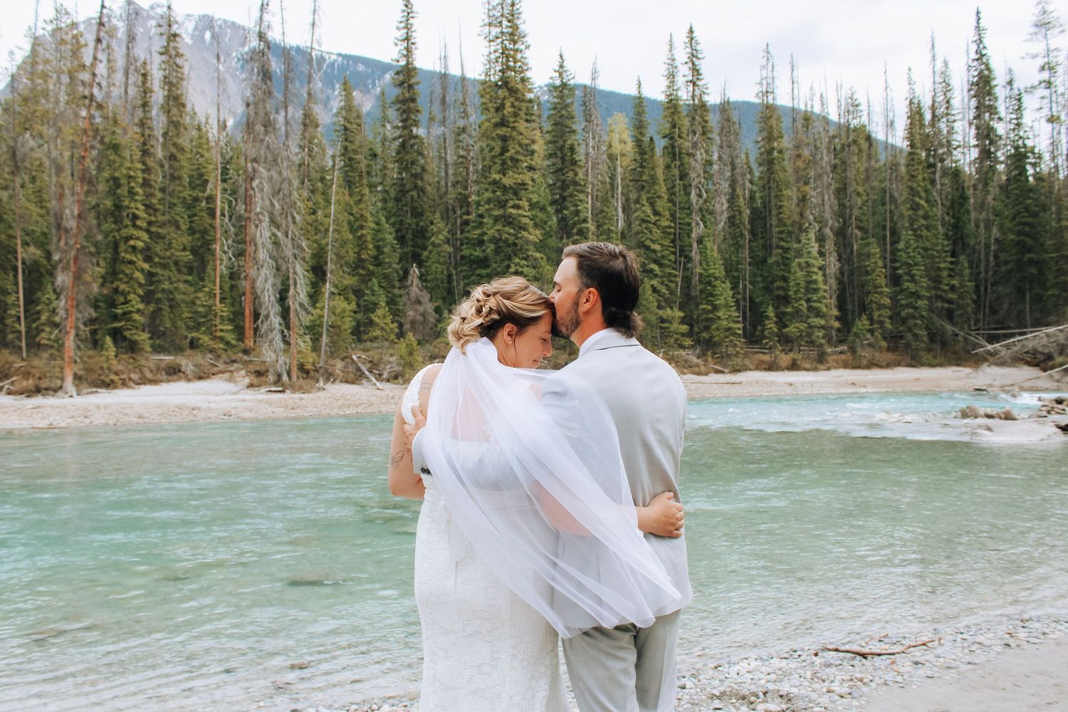 Wedding couple walking along scenic riverside with mountains and evergreens.