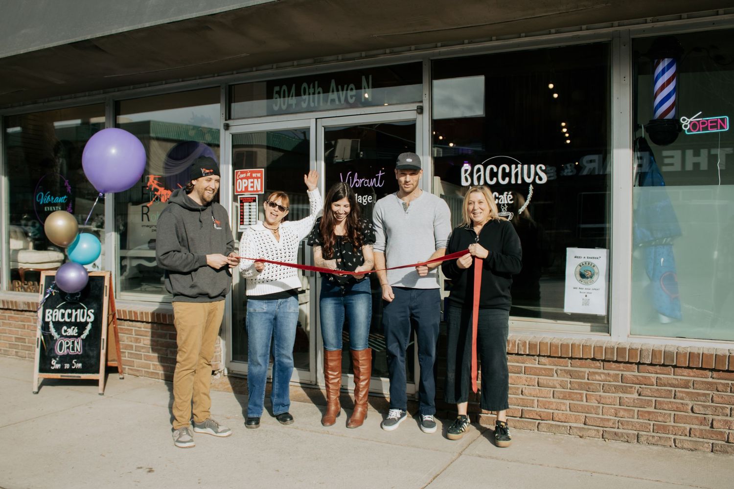 Group of people cutting ribbon at Bacchus On Elm grand opening celebration with balloons outside storefront.