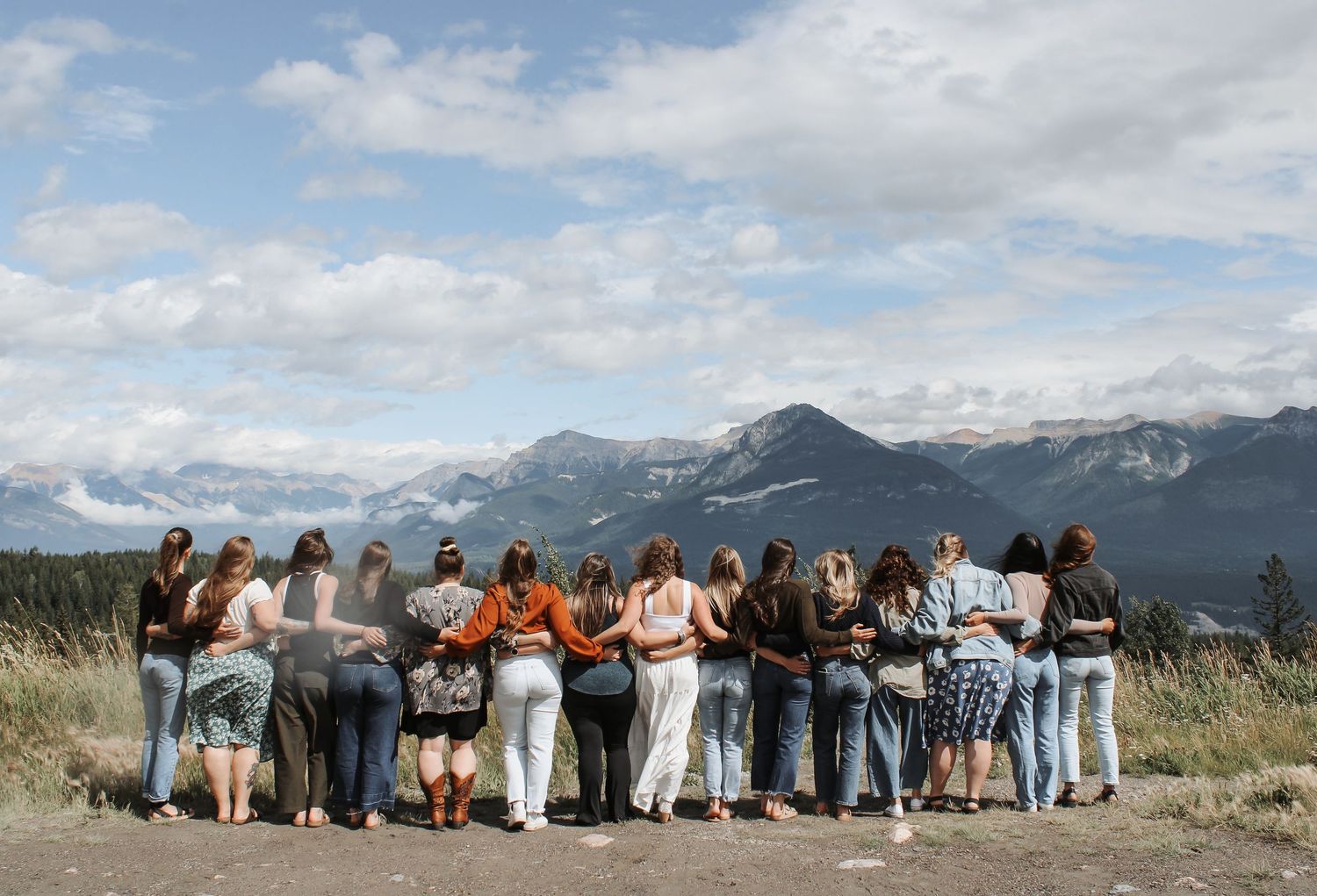 Group of women standing arm in arm overlooking a scenic mountain landscape under a cloudy sky.