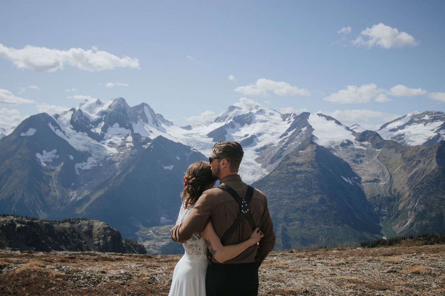 Bride in a white dress embraces a groom in suspenders on a rocky alpine ridge with snow-capped mountains and a blue sky behind them.