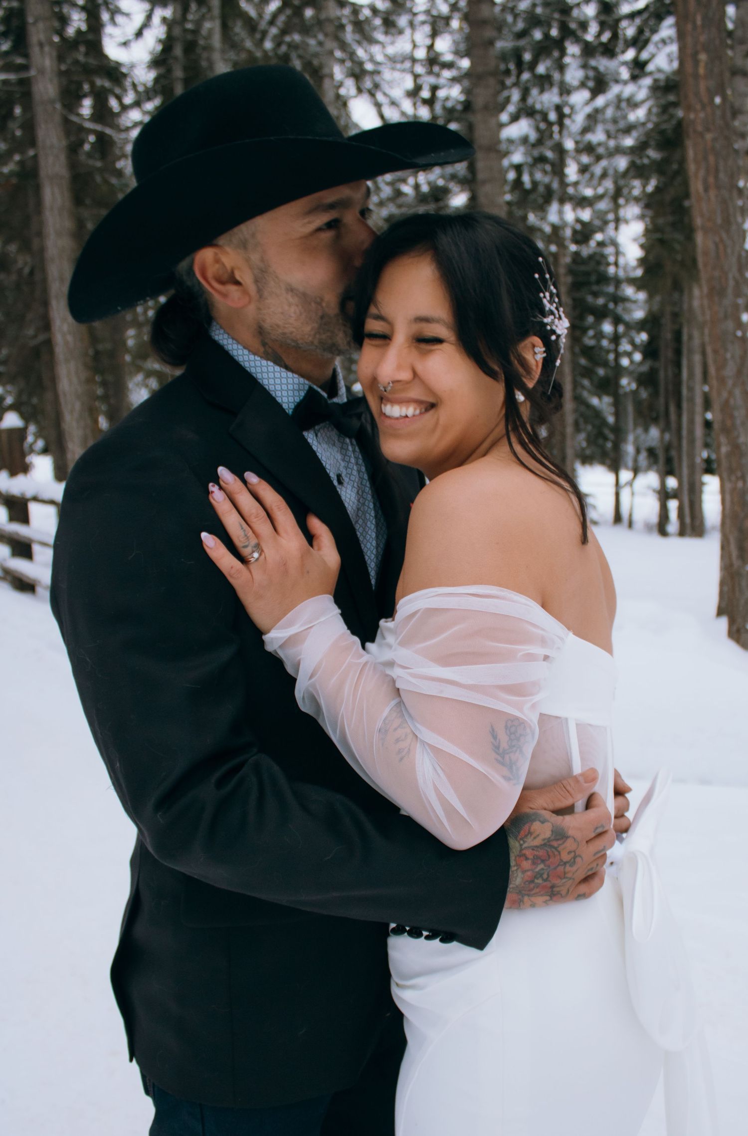 A romantic winter wedding couple embraces in a snowy forest setting wearing black and white attire.