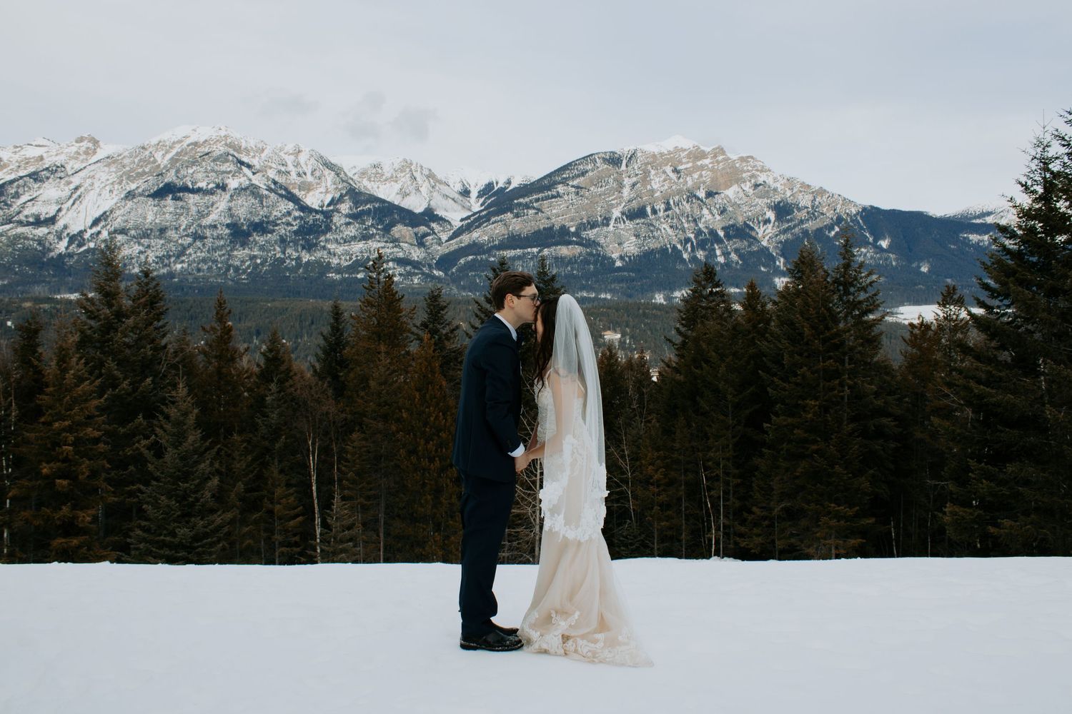 Bride in flowing white gown stands against snowy mountain backdrop during winter wedding photoshoot.