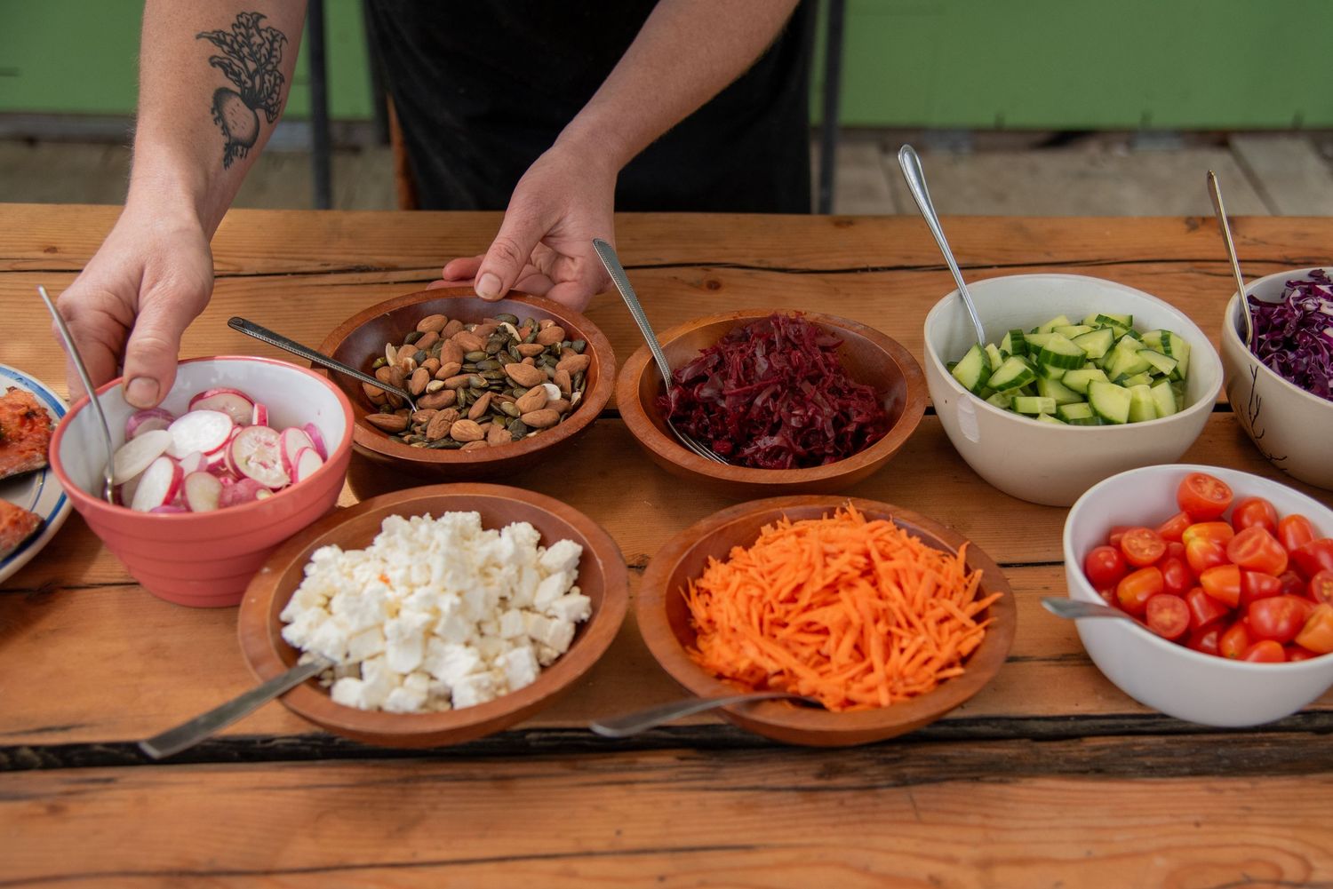 Person serving from bowls of fresh salad toppings including carrots, tomatoes, cucumber, beets, and feta on a wooden table.