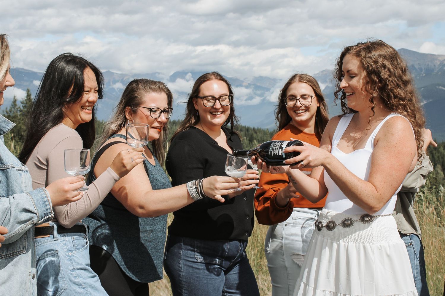 Six women smiling and toasting with wine glasses outdoors with mountain views on a sunny day.