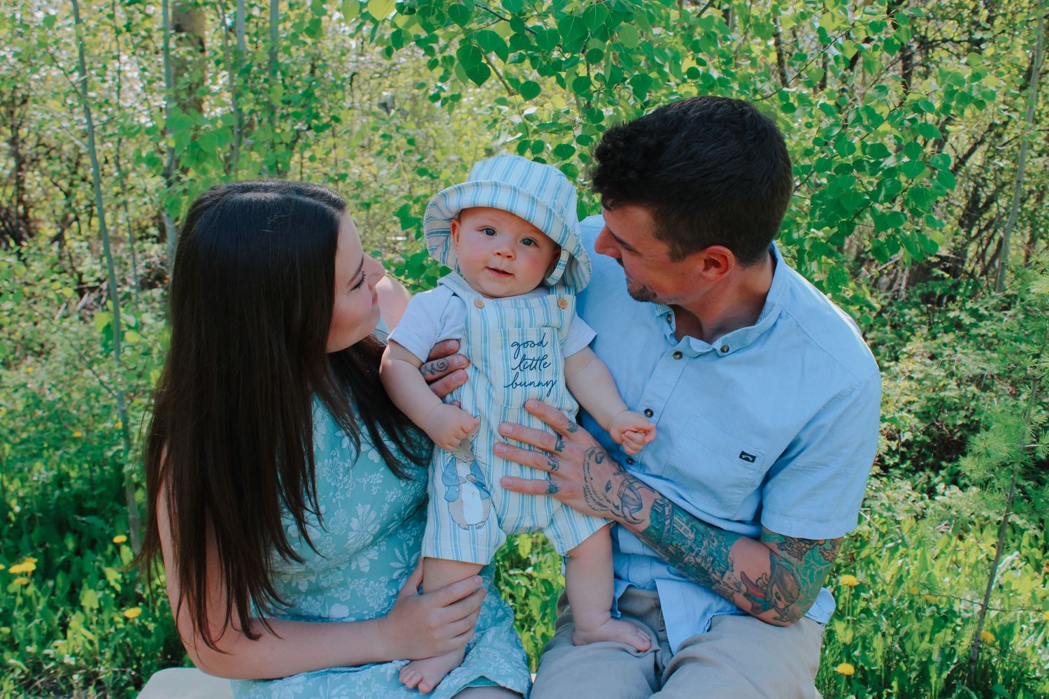 Young couple kissing baby in blue hat outdoors, surrounded by lush green trees in a family portrait.