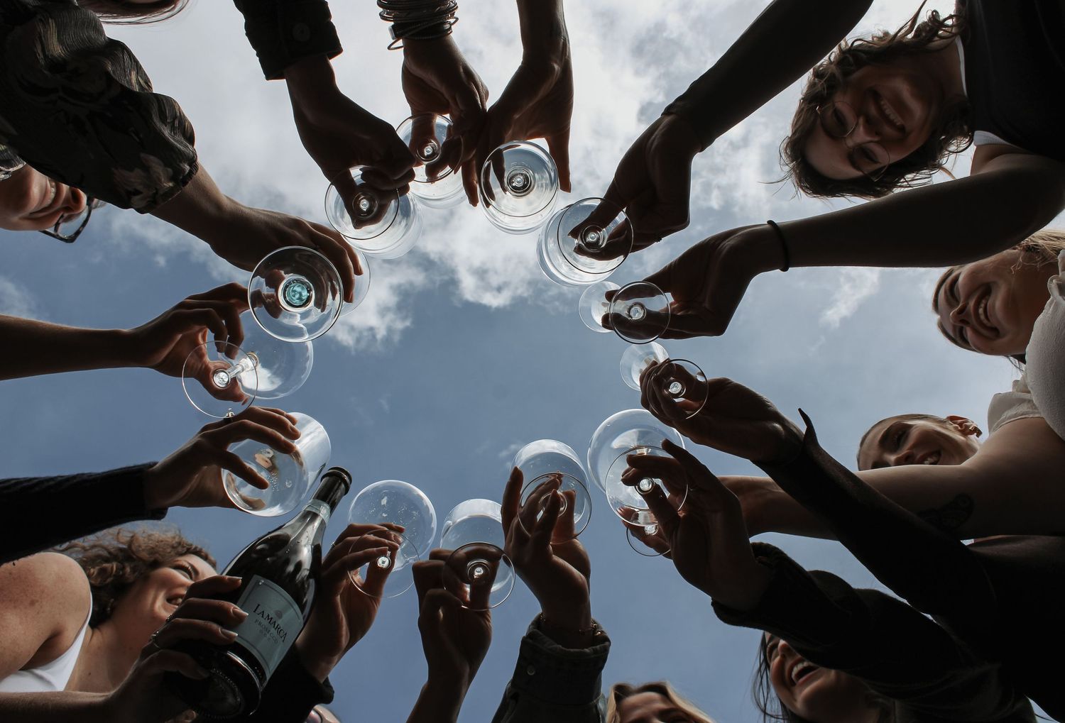 Group of people raising wine glasses in a circle toast, viewed from below against a cloudy sky.