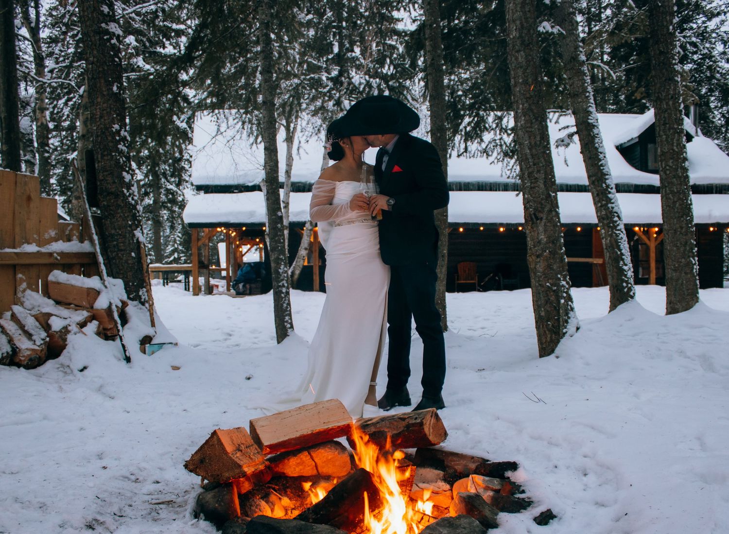 A couple stands near a glowing campfire in a snowy forest setting during a winter evening.