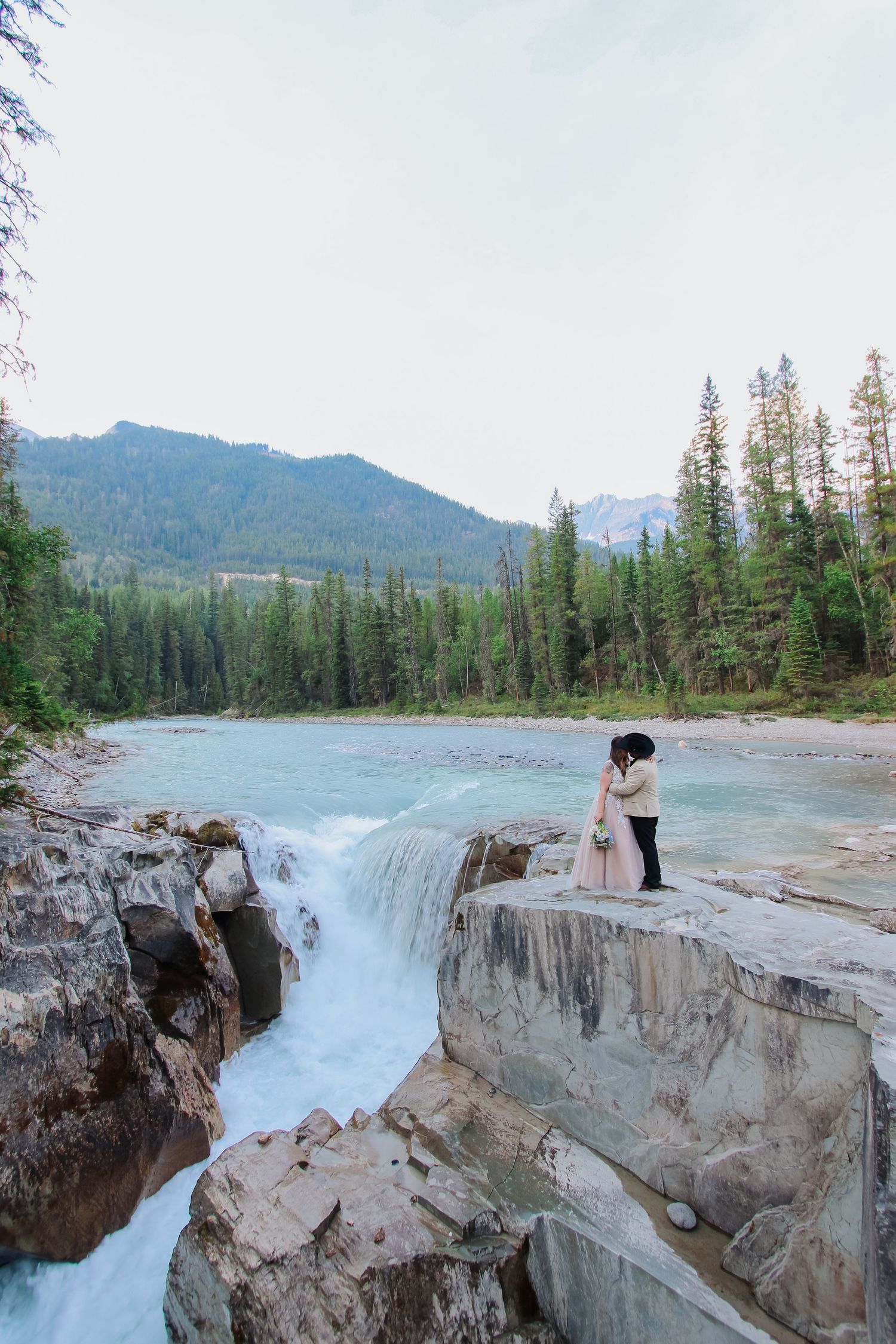 Wedding couple embracing on rocky ledge beside cascading waterfall with turquoise river and mountain forest backdrop.