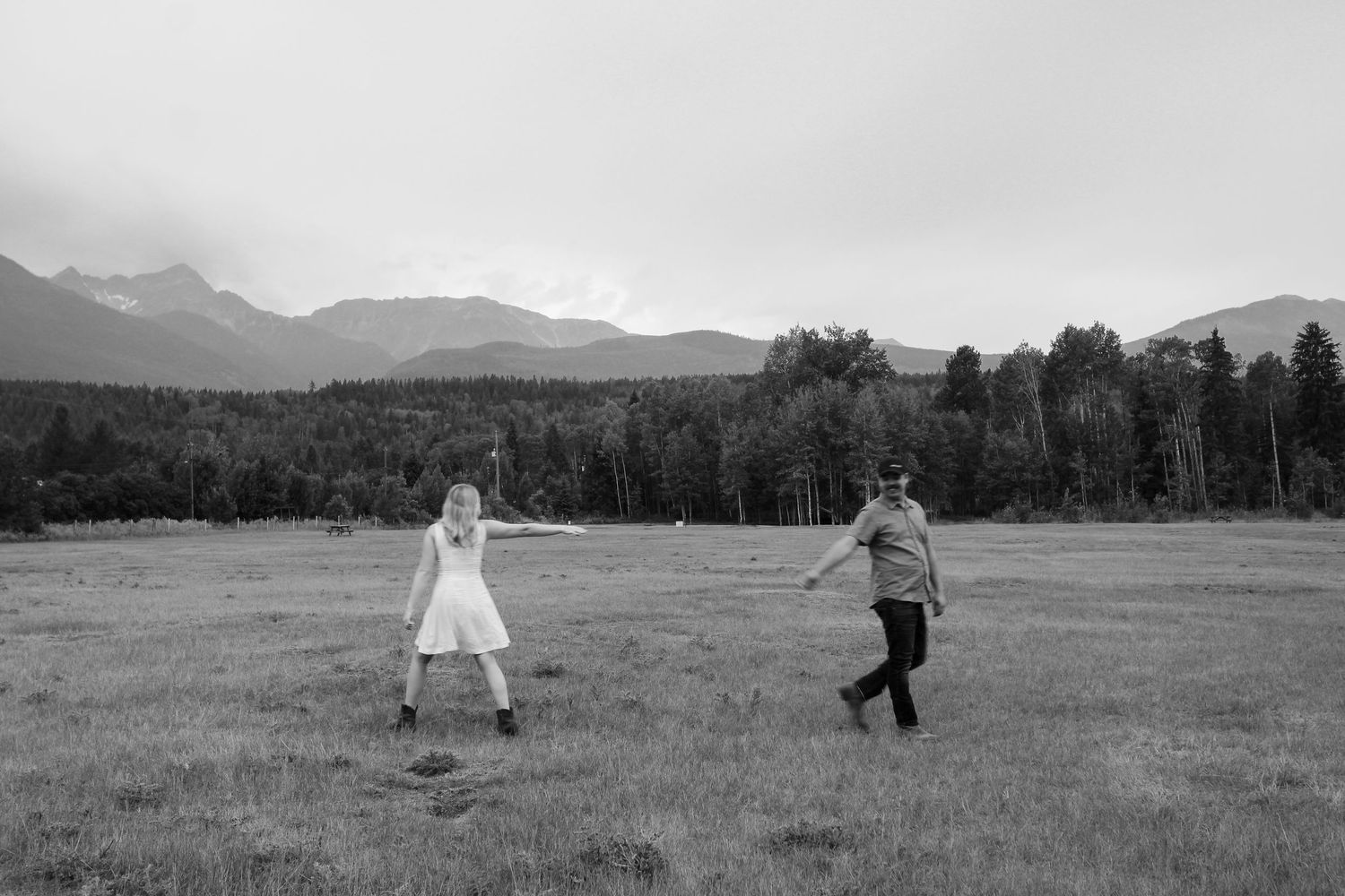 Black and white photo of a couple playfully running through a mountain meadow with forest and peaks in the background.