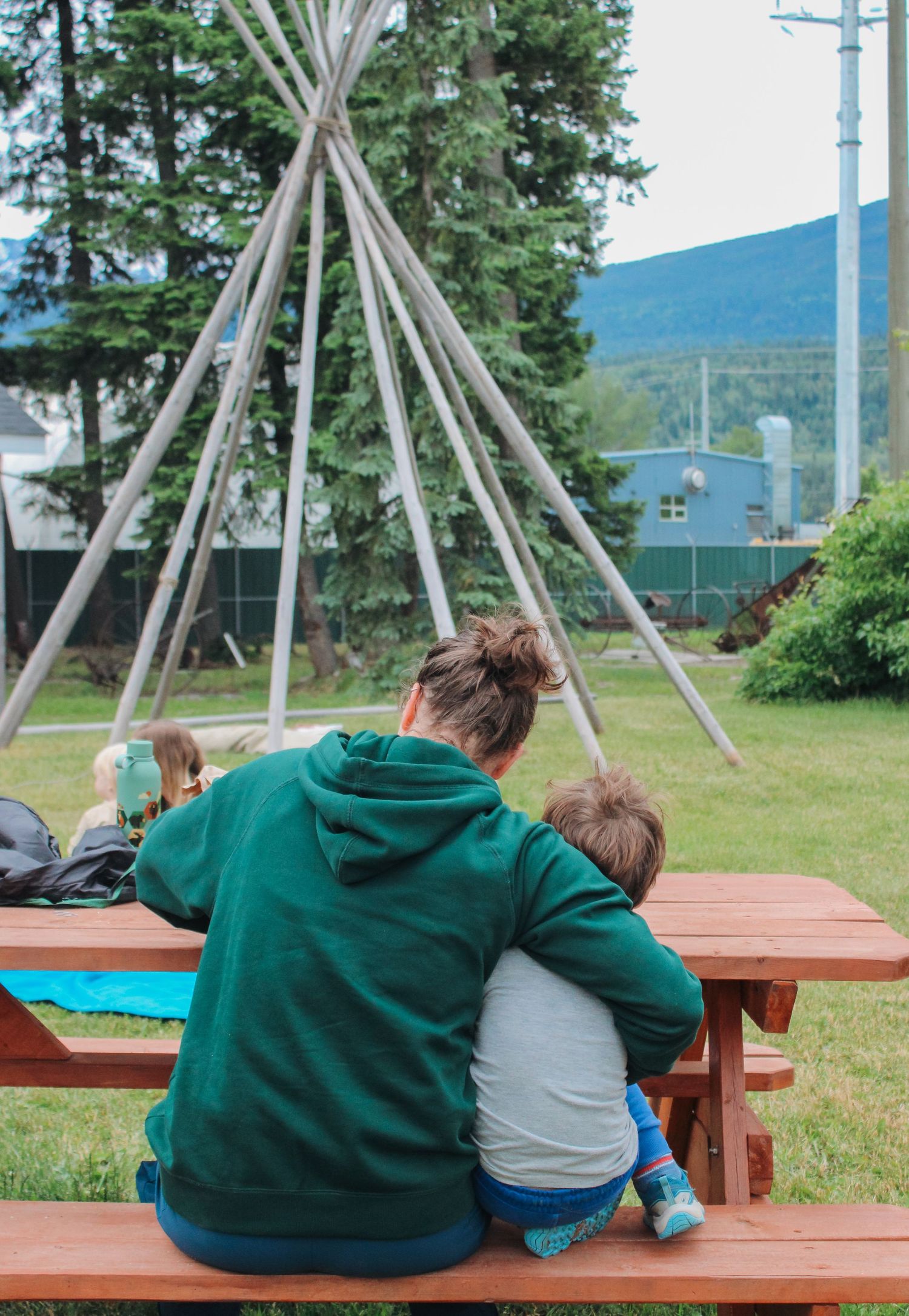 A woman in a green hoodie sits at a picnic table with a child near a teepee frame outdoors.