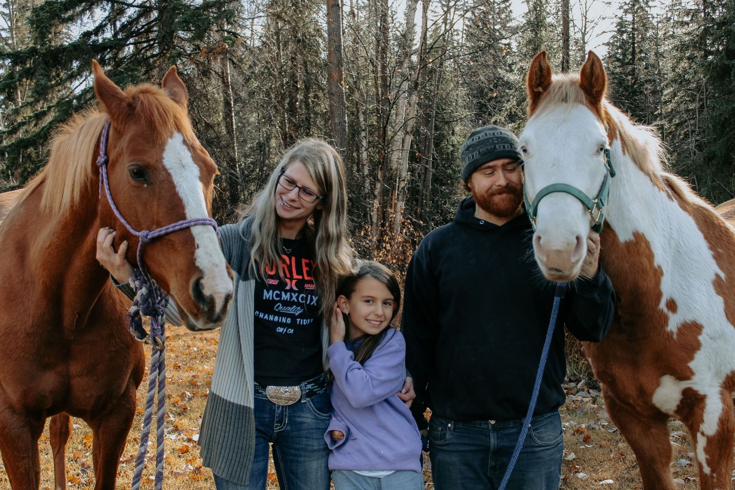 A group gathers around horses in a wooded area during golden hour.