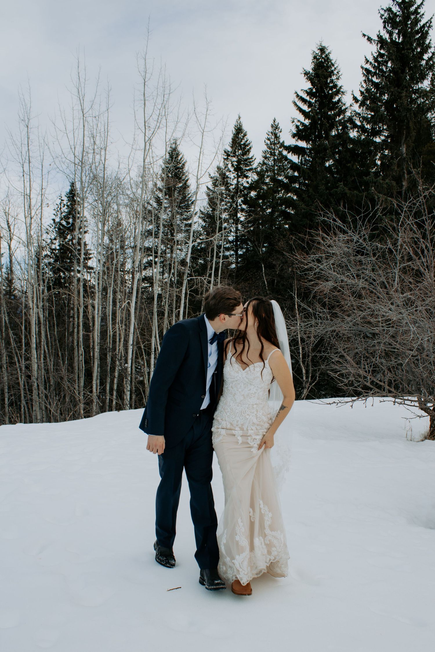 A couple shares an intimate moment in a snowy winter forest landscape wearing formal wedding attire.