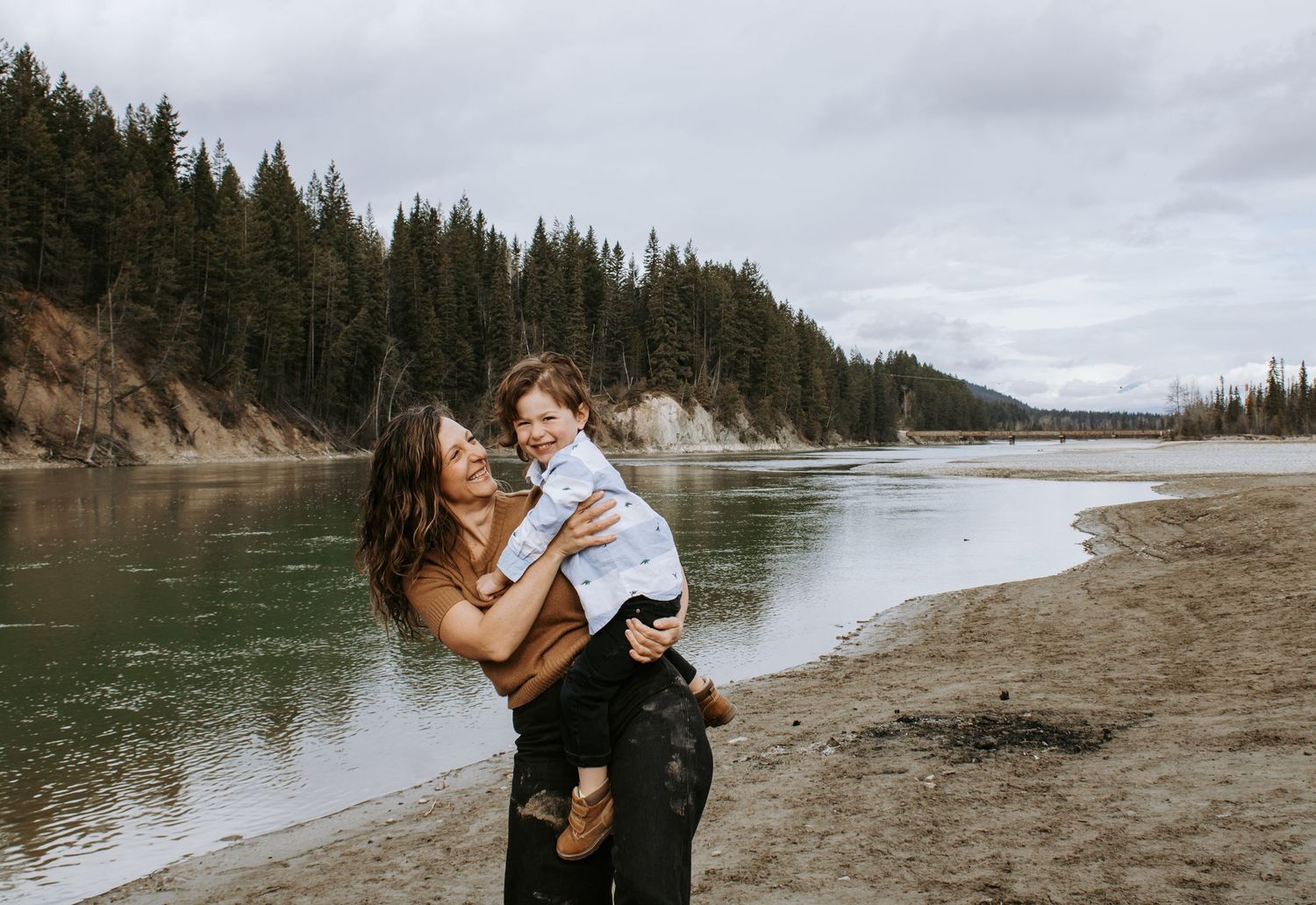 A mother joyfully lifts her toddler on a scenic river beach surrounded by evergreen trees.