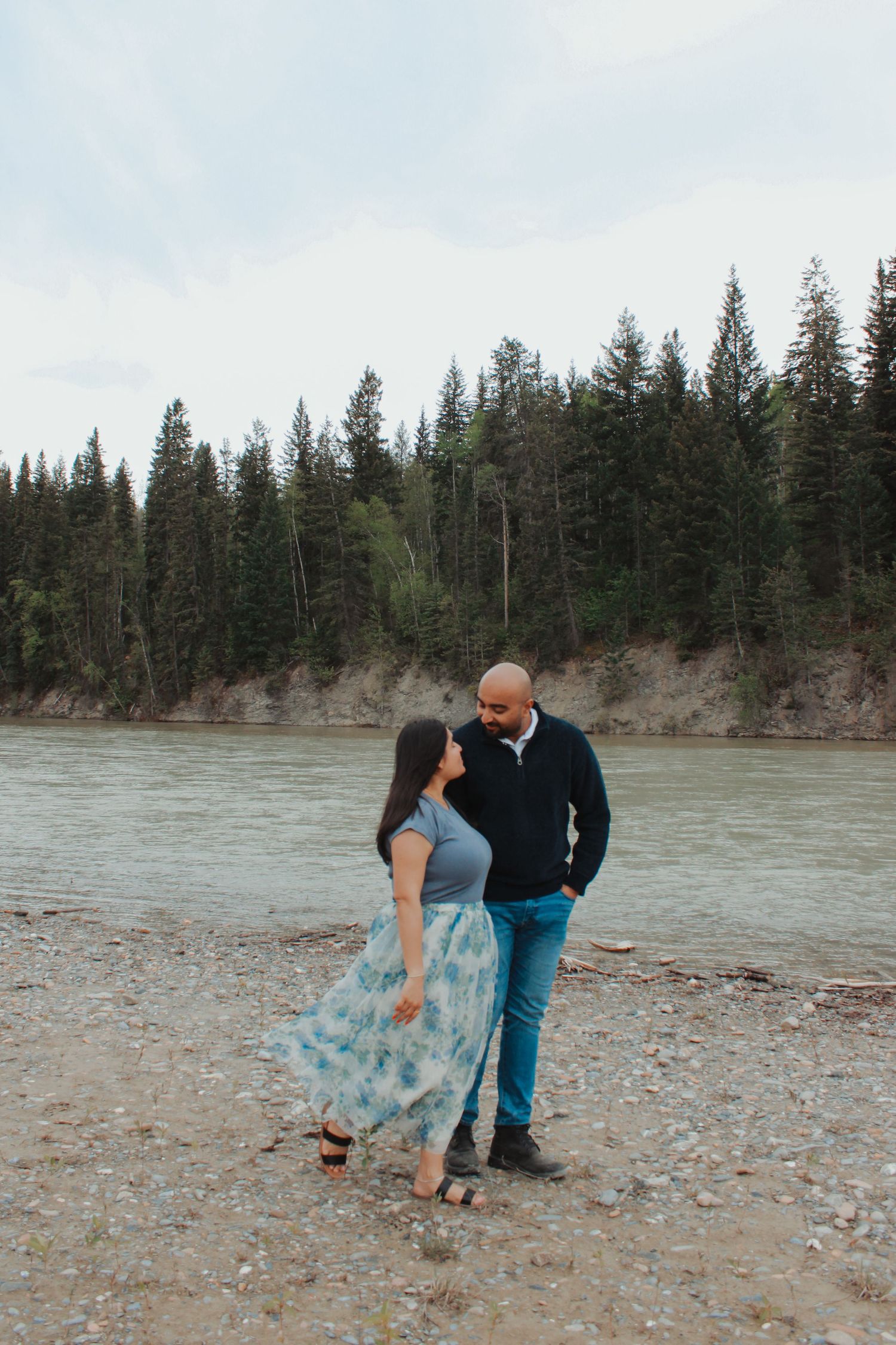 Couple sharing a romantic moment by a river with evergreen forest backdrop on a cloudy day.
