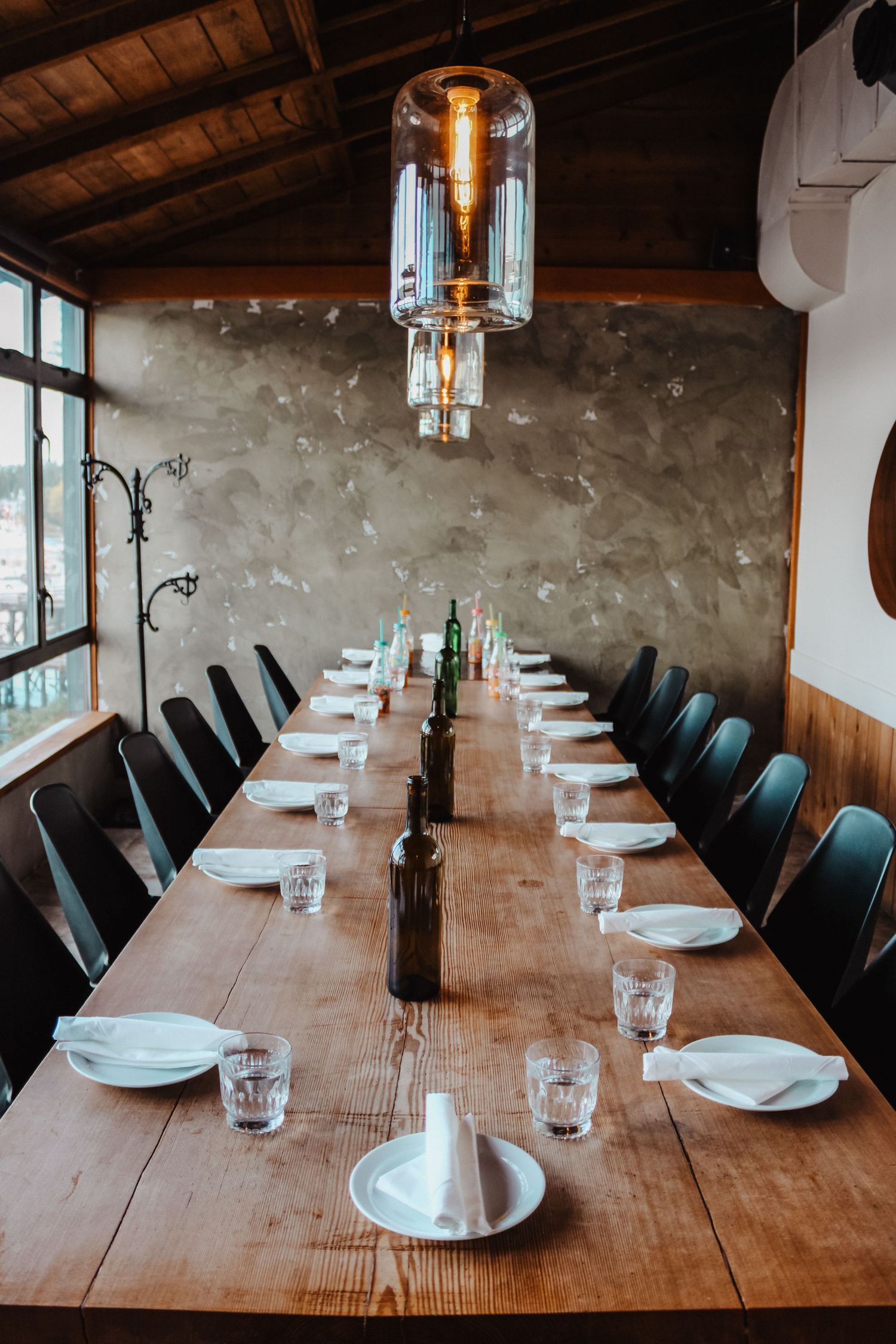 Long wooden dining table set with white plates and glasses in a rustic room with wooden beams.
