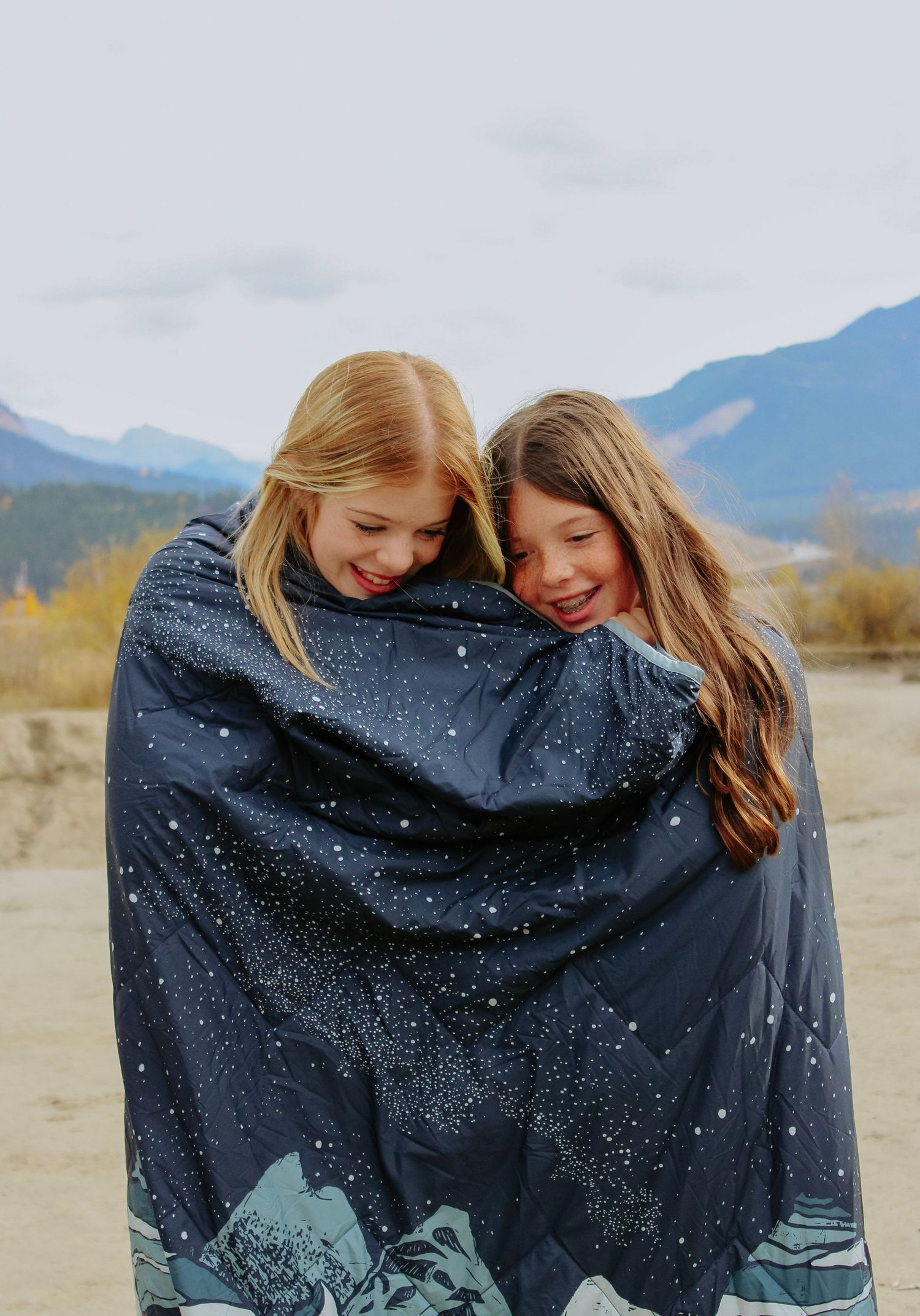 Two people share a cozy moment under a starry blanket in a mountainous landscape.