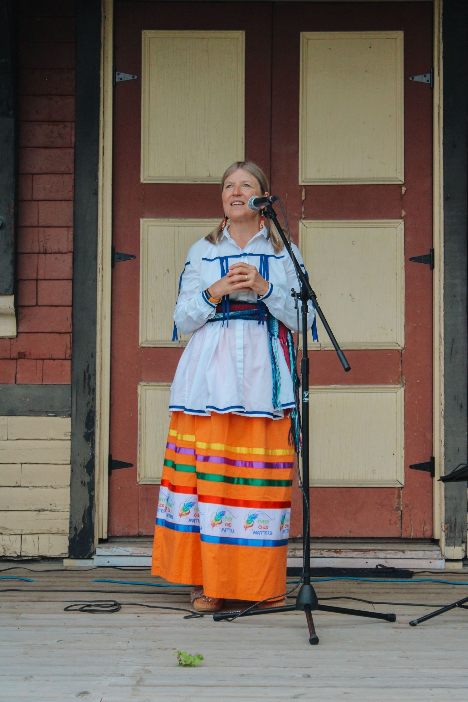 Woman in colorful traditional folk costume speaking at outdoor microphone stand on wooden stage.