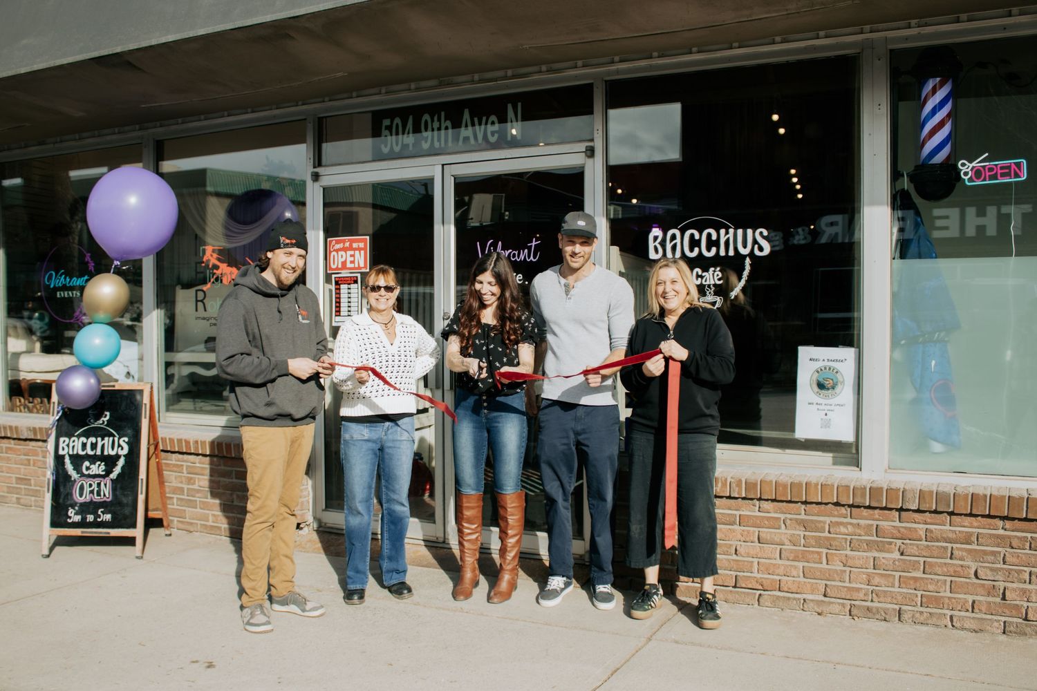 Group of people cutting a ribbon at the grand opening of Bacchus bar on 6th Ave, with balloons and signage outside.
