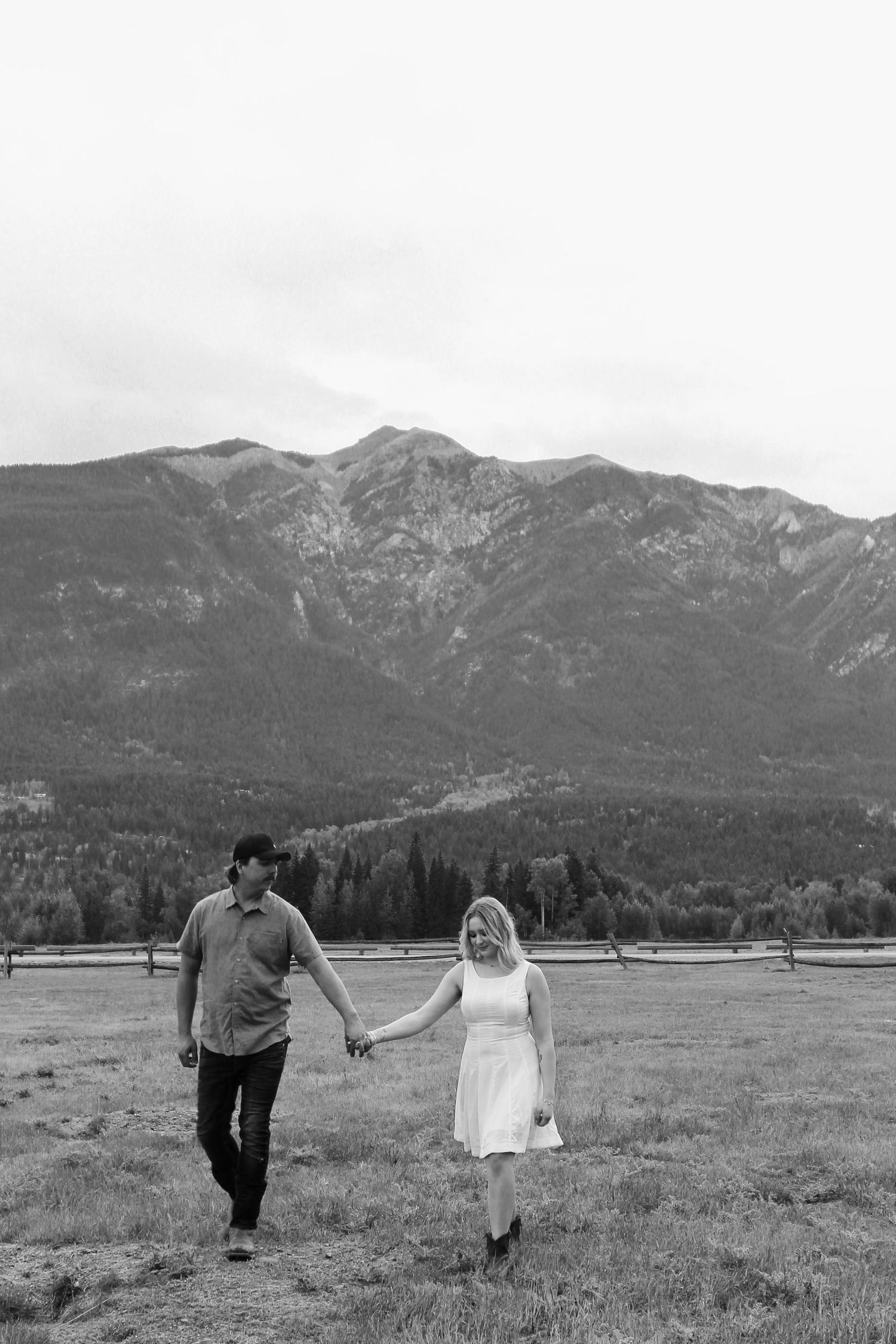 Black and white photo of a couple holding hands walking in a mountain meadow with forested peaks in the background.