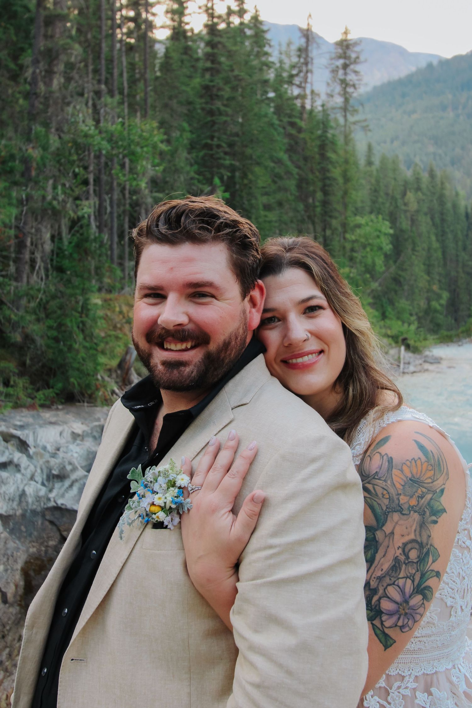 Smiling bride with floral tattoo sleeve embraces groom in tan blazer with boutonniere, posing outdoors near forest and mountains.