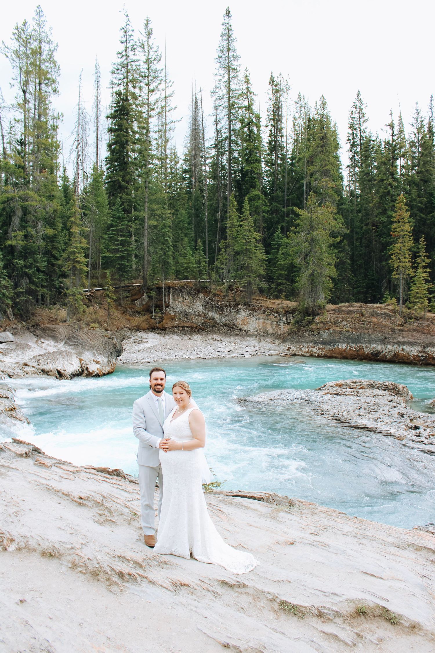 Couple poses along turquoise river with evergreen forest backdrop in natural mountain setting.