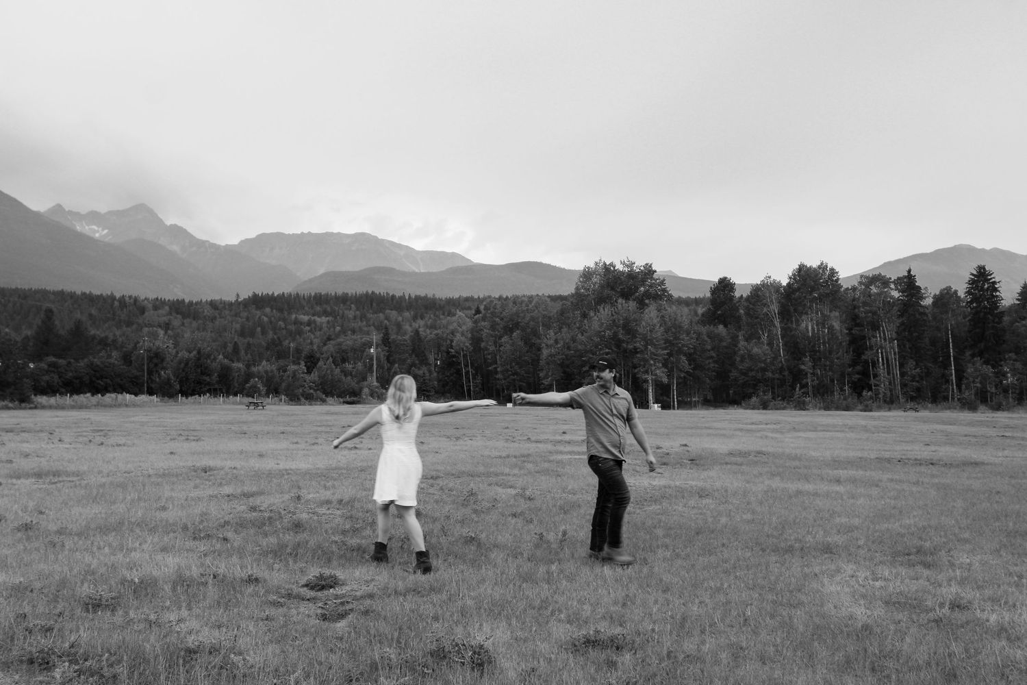 Black and white photo of a couple holding hands in a mountain meadow with forest and peaks in the background.