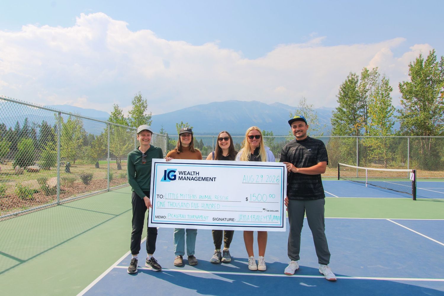 Five people holding a large IG Wealth Management donation check on an outdoor tennis court with mountain views.