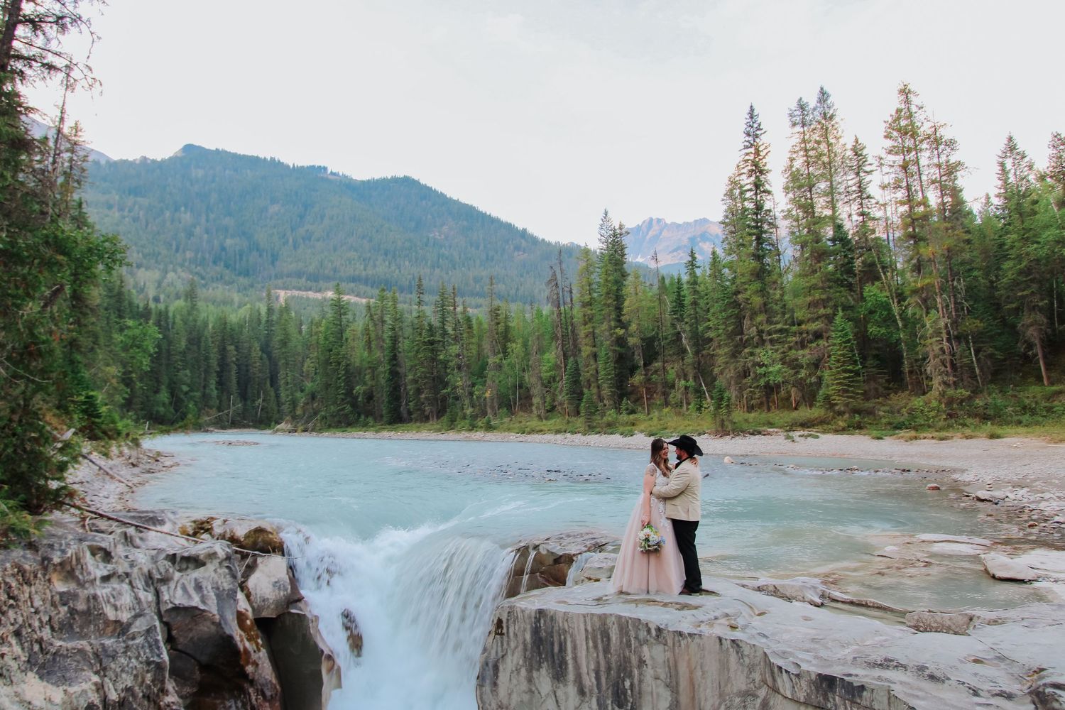 Couple embracing on rocky waterfall edge with turquoise river, pine trees, and mountain backdrop in Banff National Park.