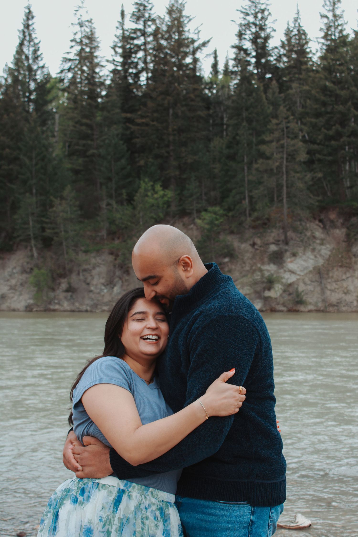Couple embracing and laughing by a river with pine trees in the background during an outdoor engagement session.