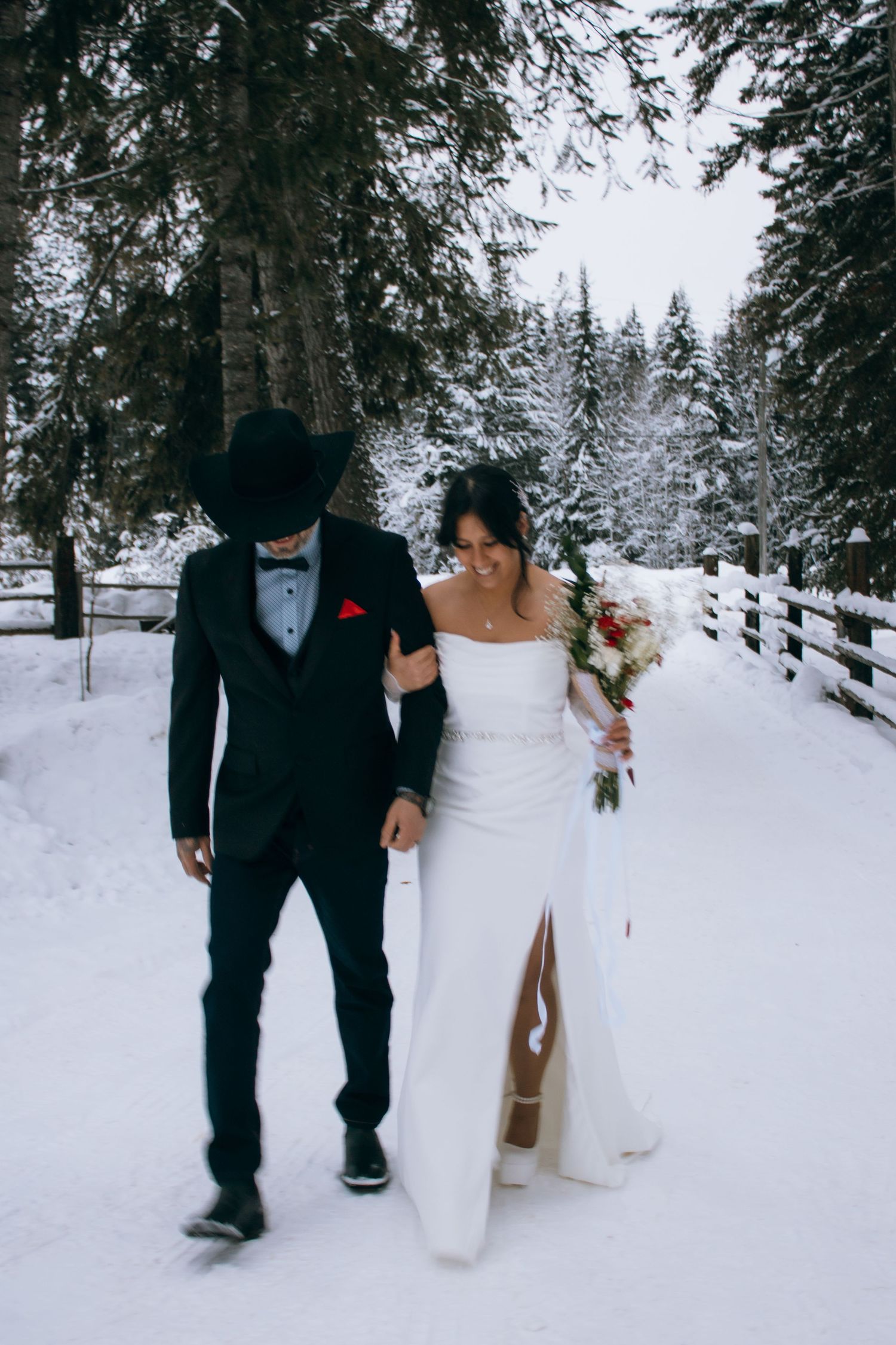 A couple walks together through a snowy winter forest path during their wedding celebration.