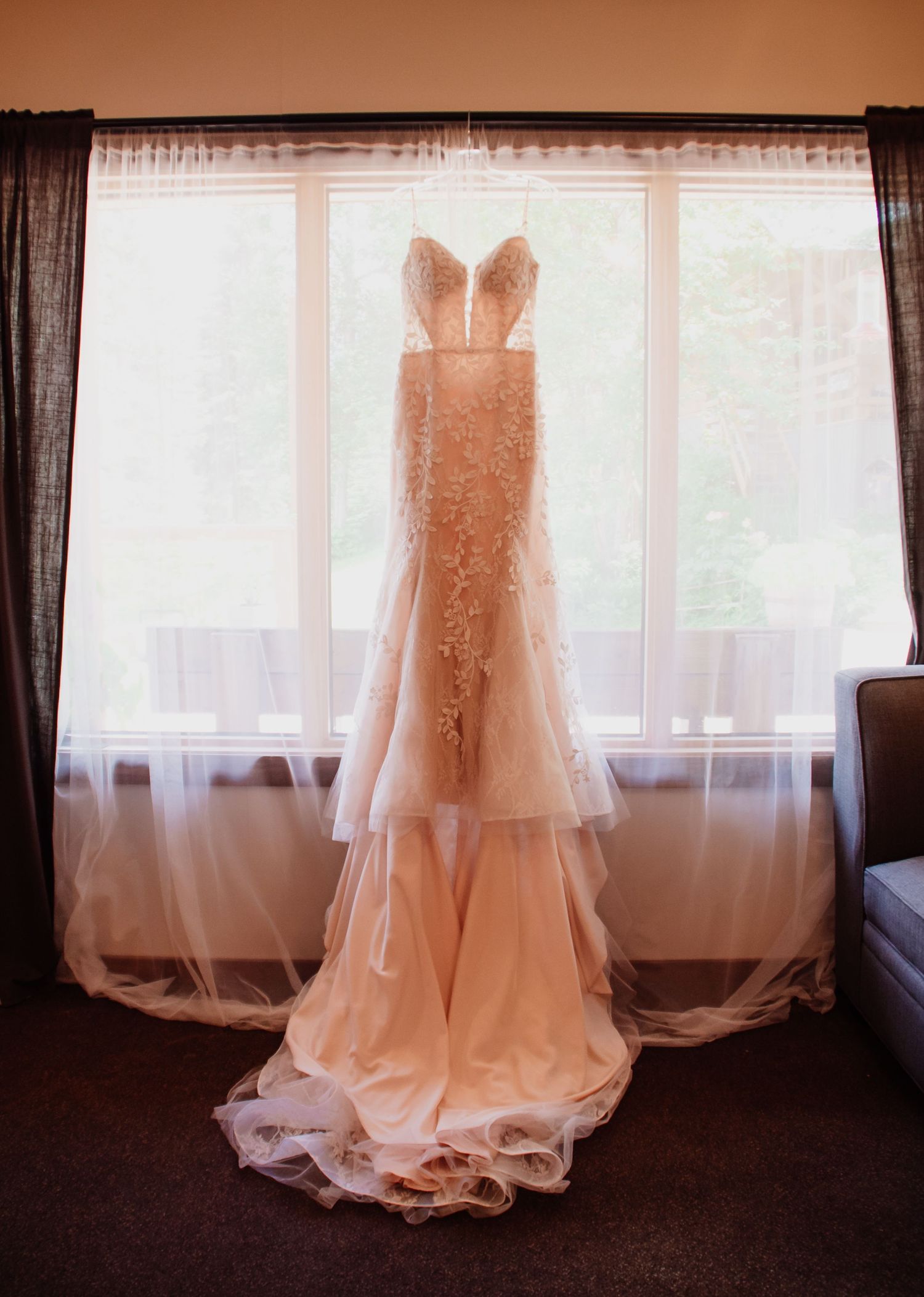 Elegant blush mermaid wedding gown with lace appliques and tiered tulle skirt hanging in a sunlit window.