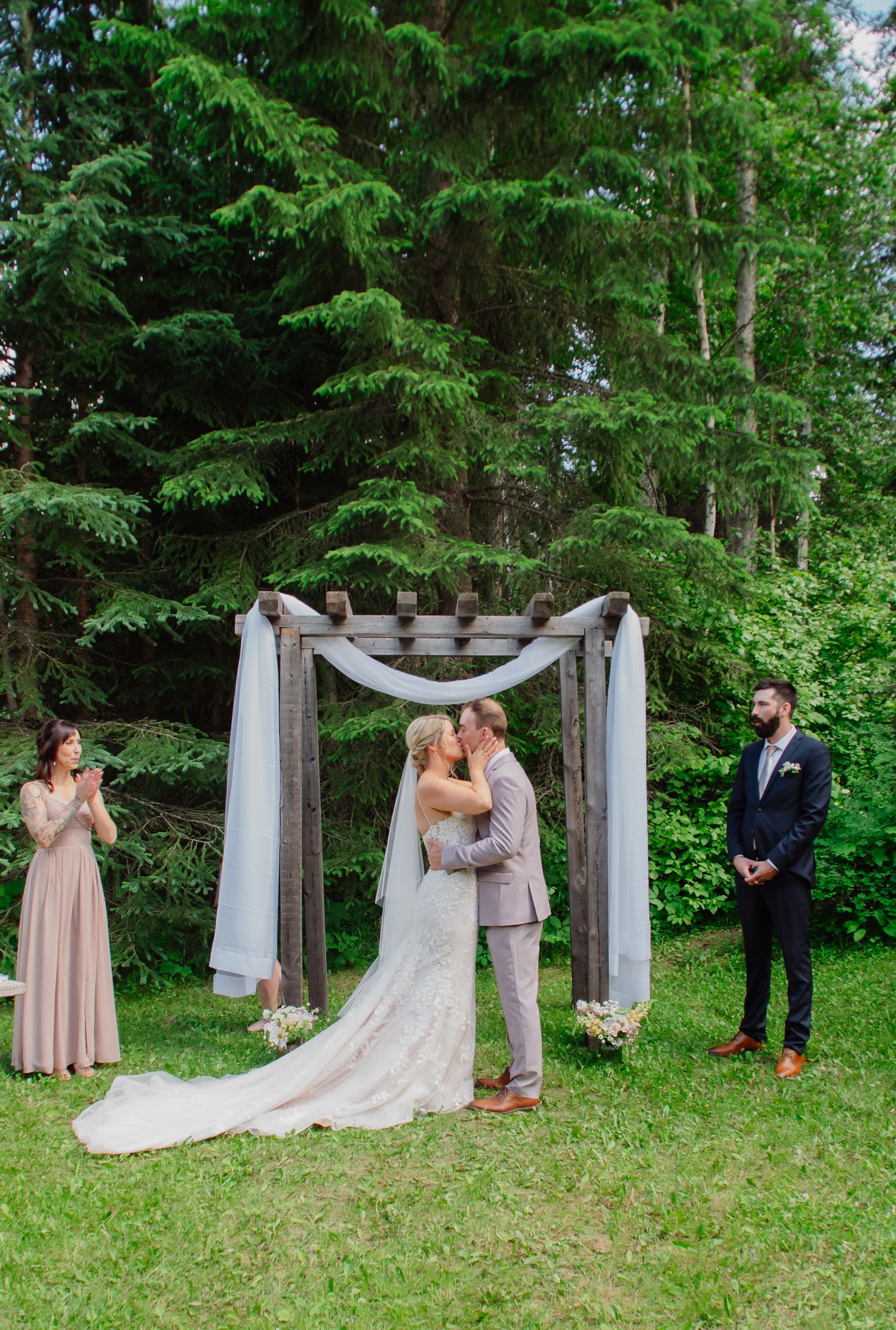 Bride and groom share first kiss under rustic wooden arch draped with white fabric at outdoor forest wedding ceremony.