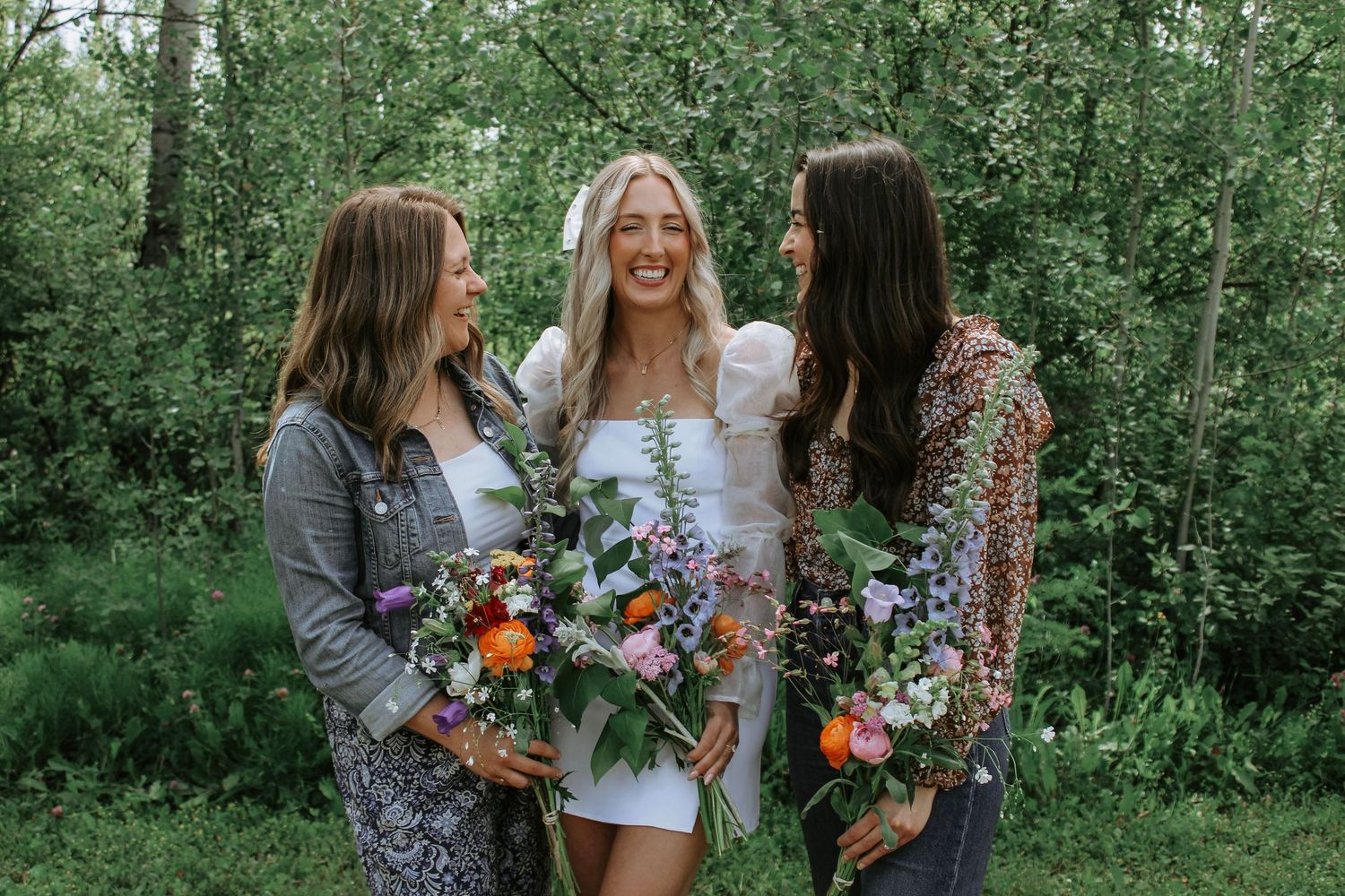 Three women smiling outdoors holding colorful flower bouquets, with lush green trees in the background.