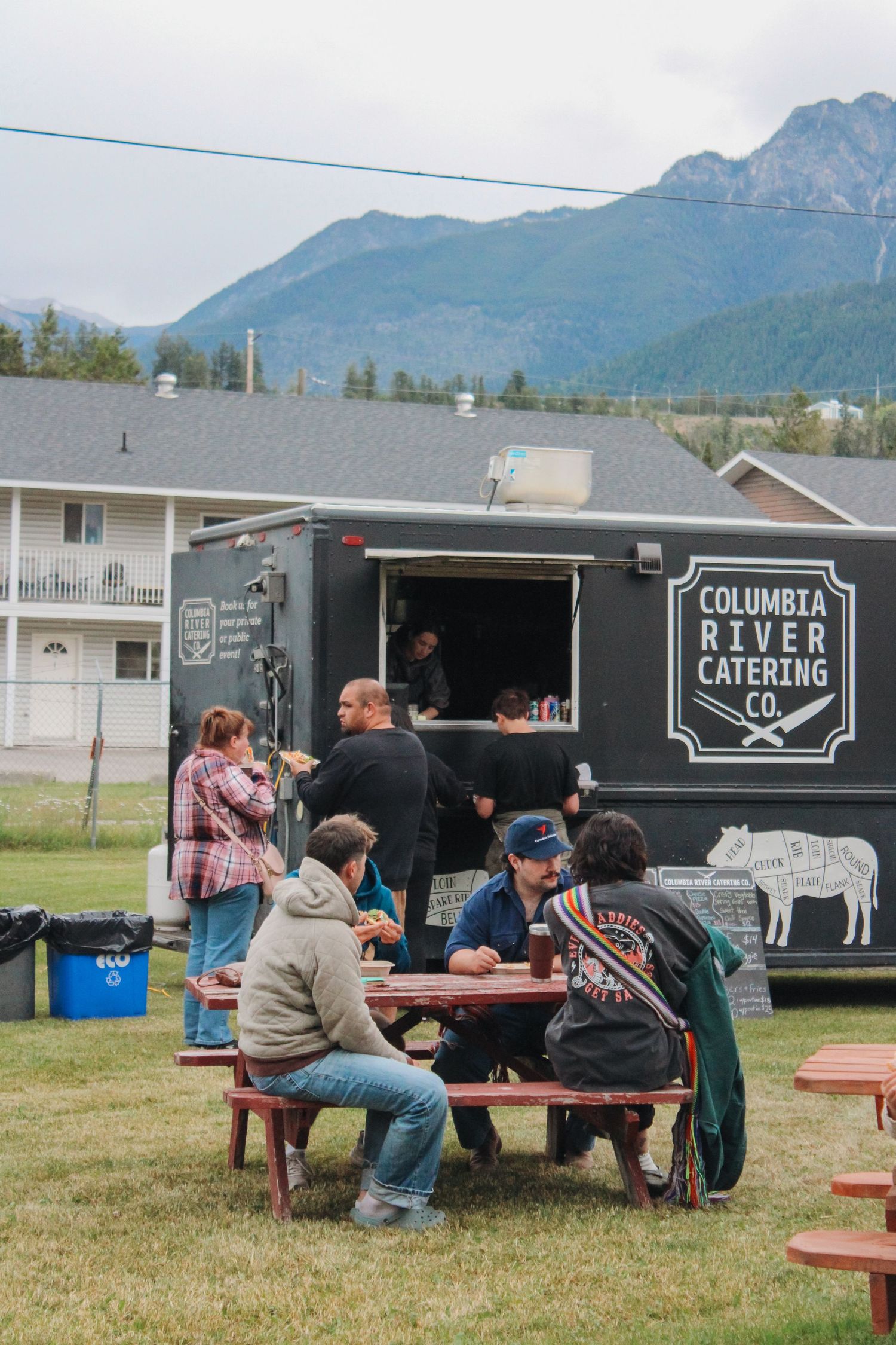 People dining at picnic tables outside Columbia River Catering Co. food truck with mountain backdrop.
