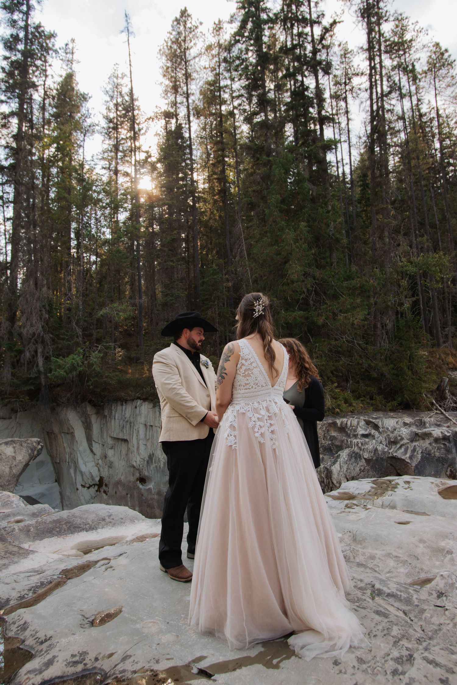 Couple exchanging vows during outdoor wedding ceremony on rocky cliff surrounded by tall pine trees at sunset.