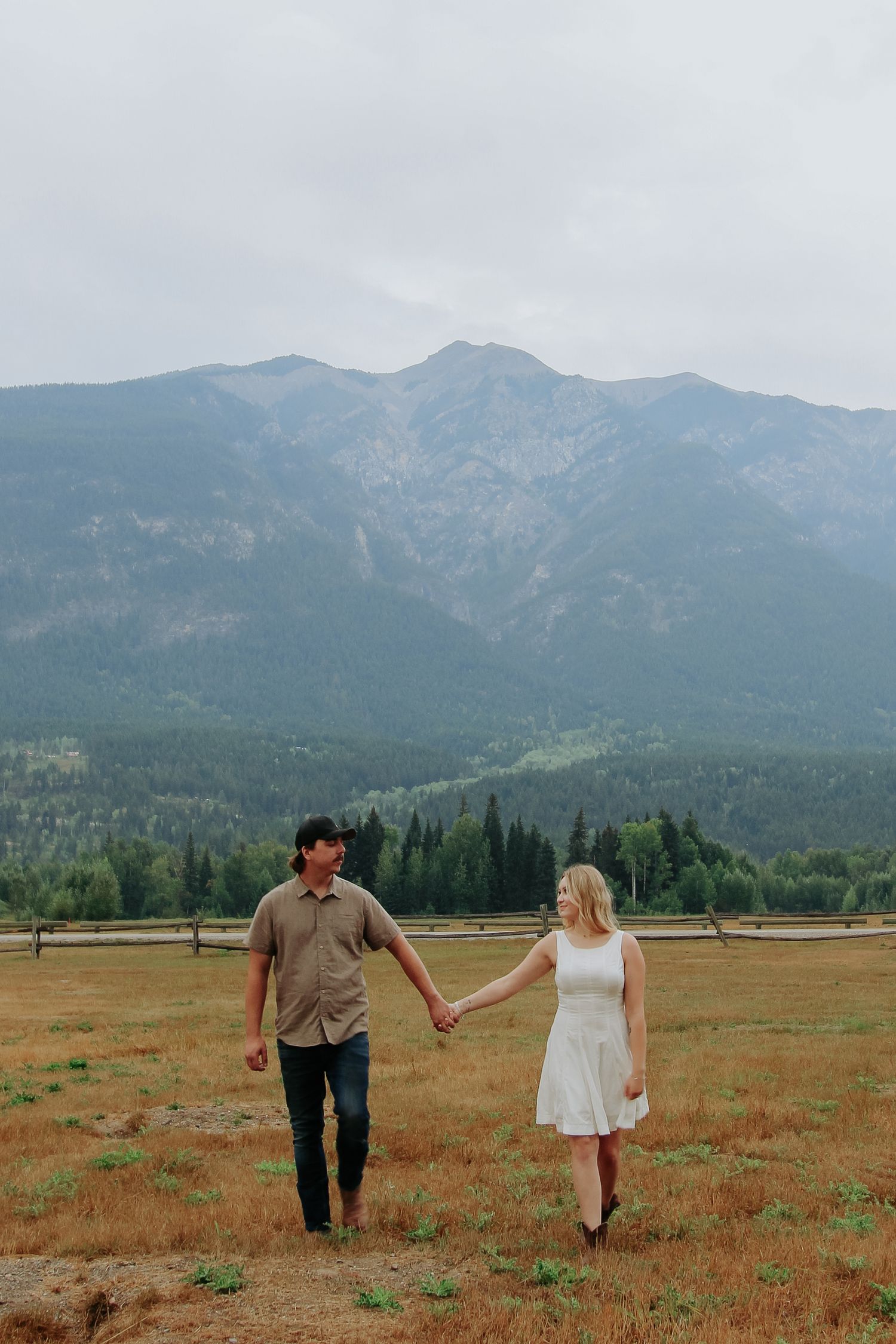 Couple holds hands walking in mountain meadow, woman in white dress, man in casual shirt, scenic peaks behind.