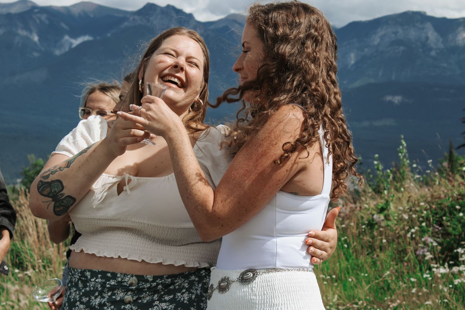 Two women laughing and embracing outdoors with mountain scenery, one holding a baby, wearing summer outfits.
