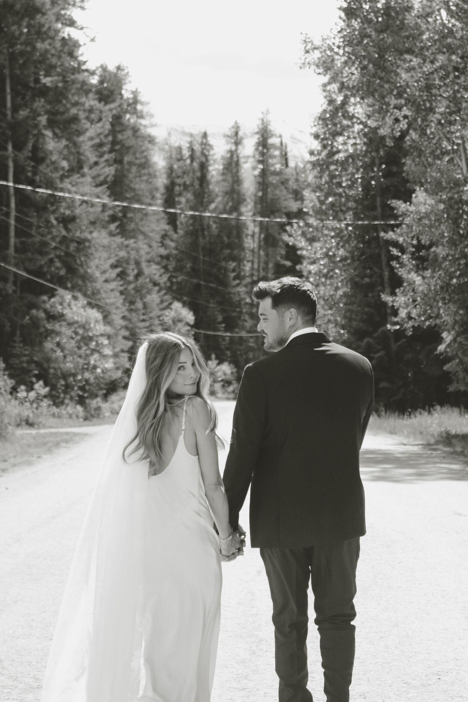 Black and white photo of a bride and groom holding hands walking down a tree-lined road, bride glancing back.
