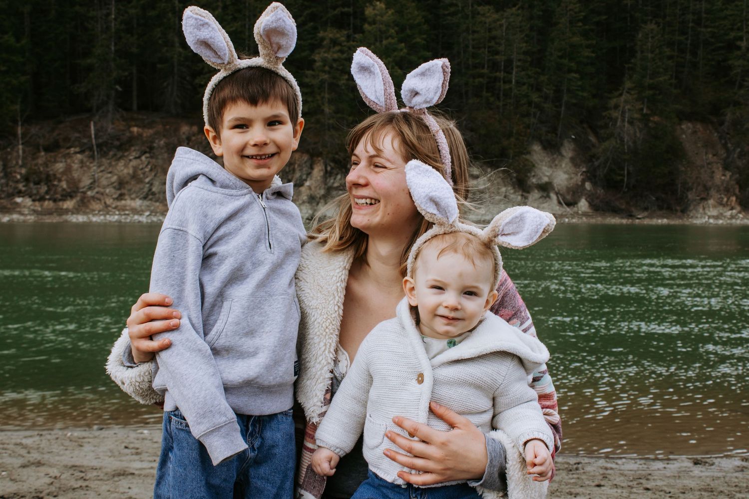 A mother and two children wearing bunny ears pose by a lake with forest backdrop for Easter photos.