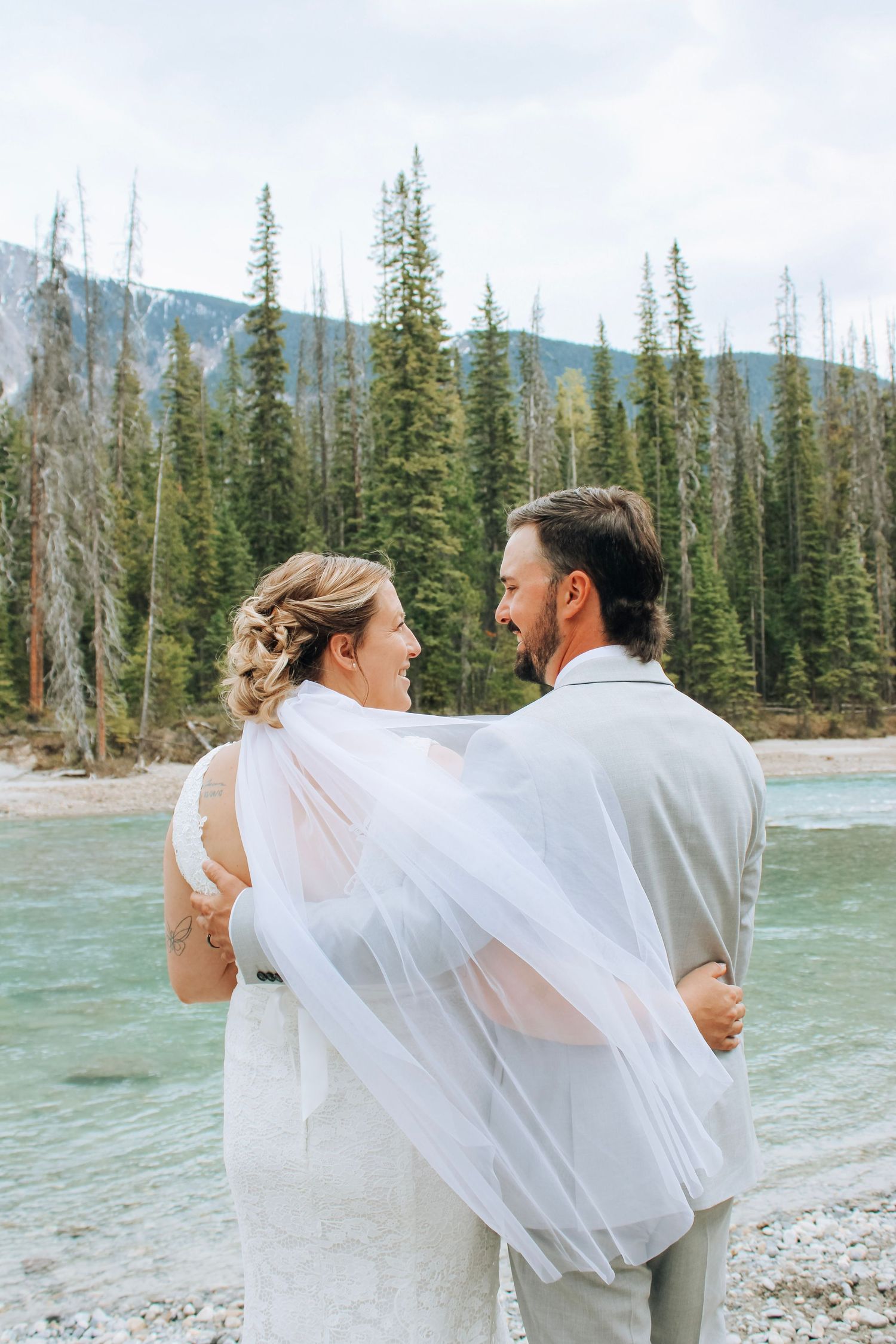 Newlyweds embracing by turquoise river with mountain backdrop and flowing veil.