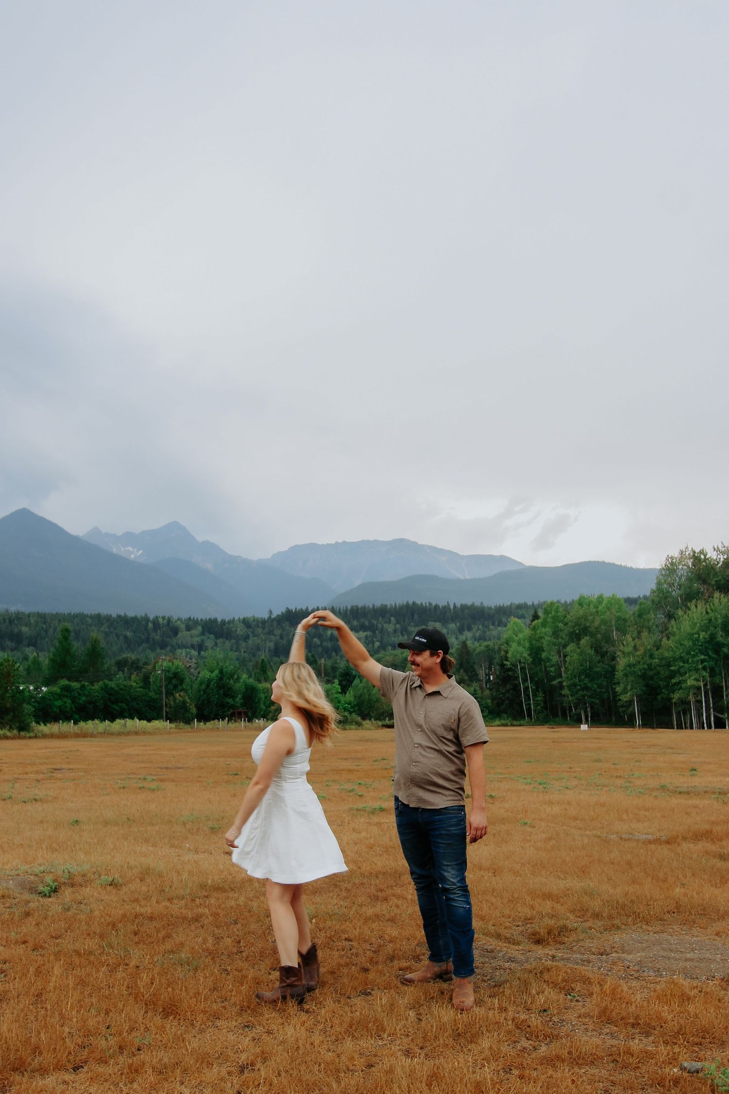 Couple dancing in a golden field with mountain and forest backdrop under cloudy skies.