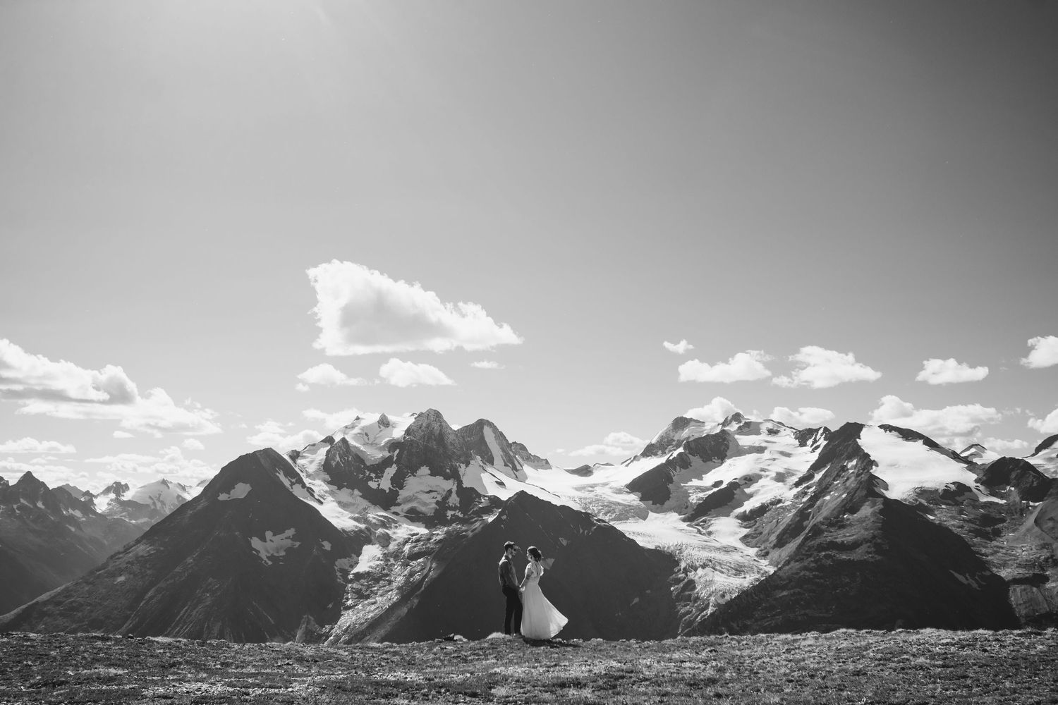 Bride and groom embrace on a mountaintop in a black and white wedding photo with rugged snow-capped peaks and clouds behind them.